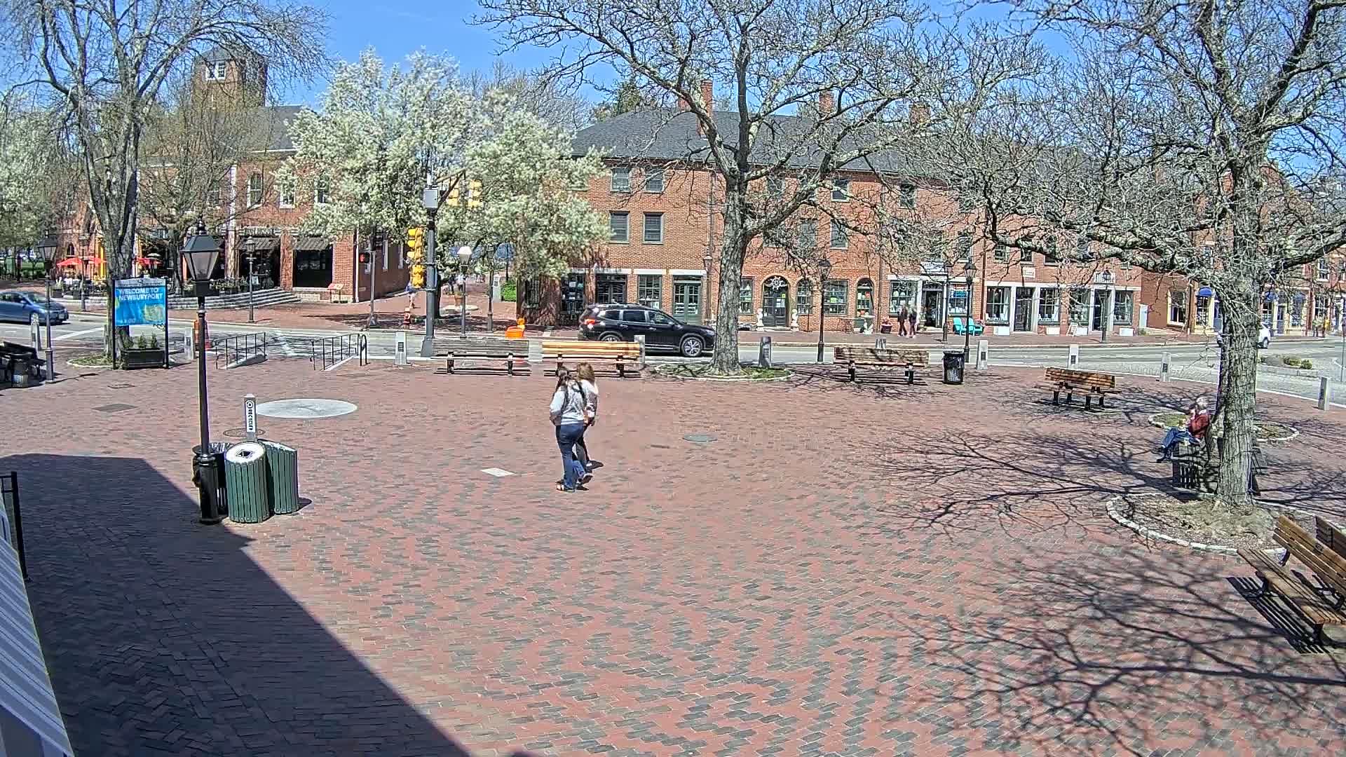 A brick plaza with benches and several people, surrounded by brick buildings and leafless trees under a sunny sky.