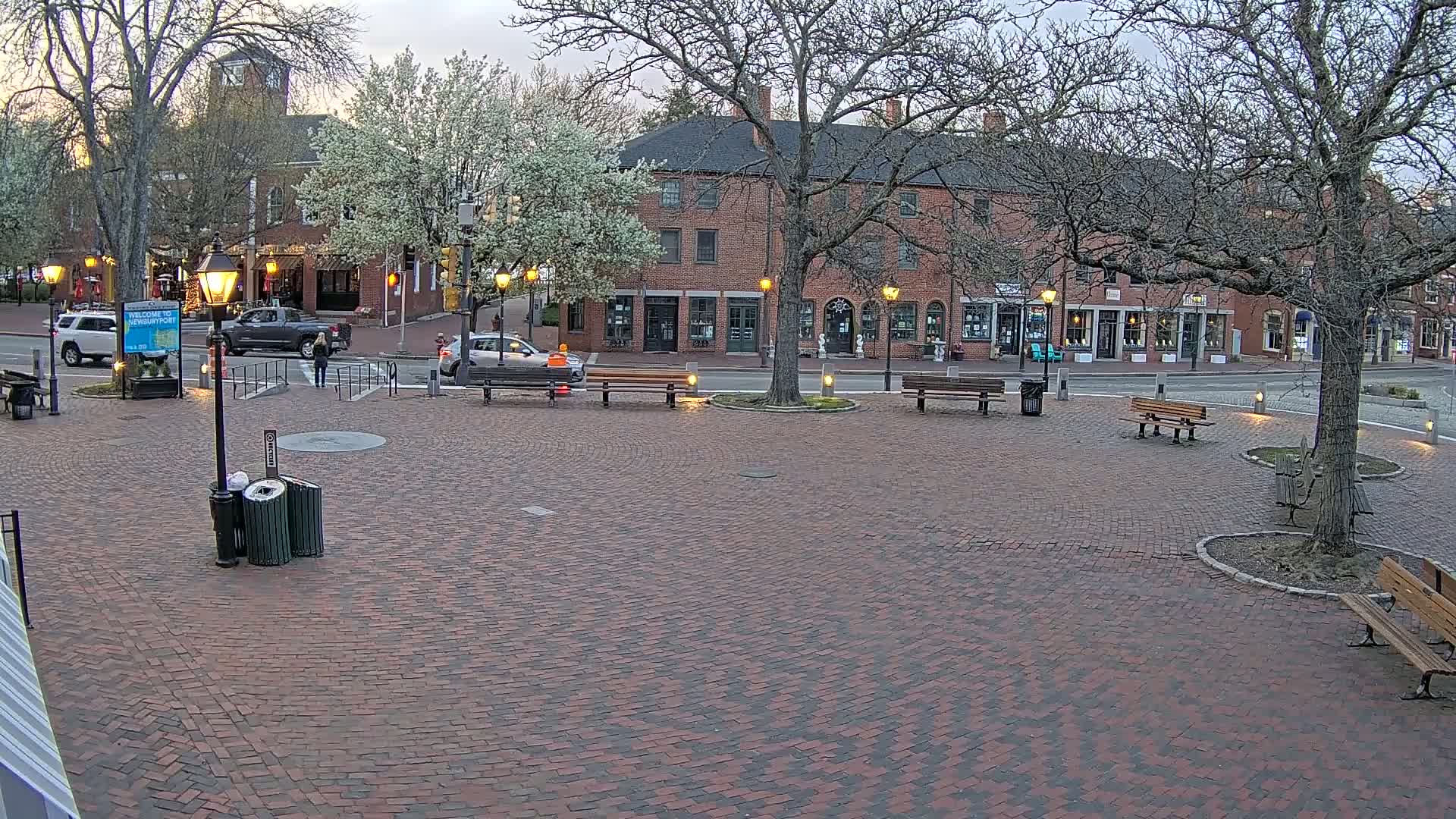 A brick plaza with several park benches and lampposts sits in front of a row of brick buildings under a cloudy sky.