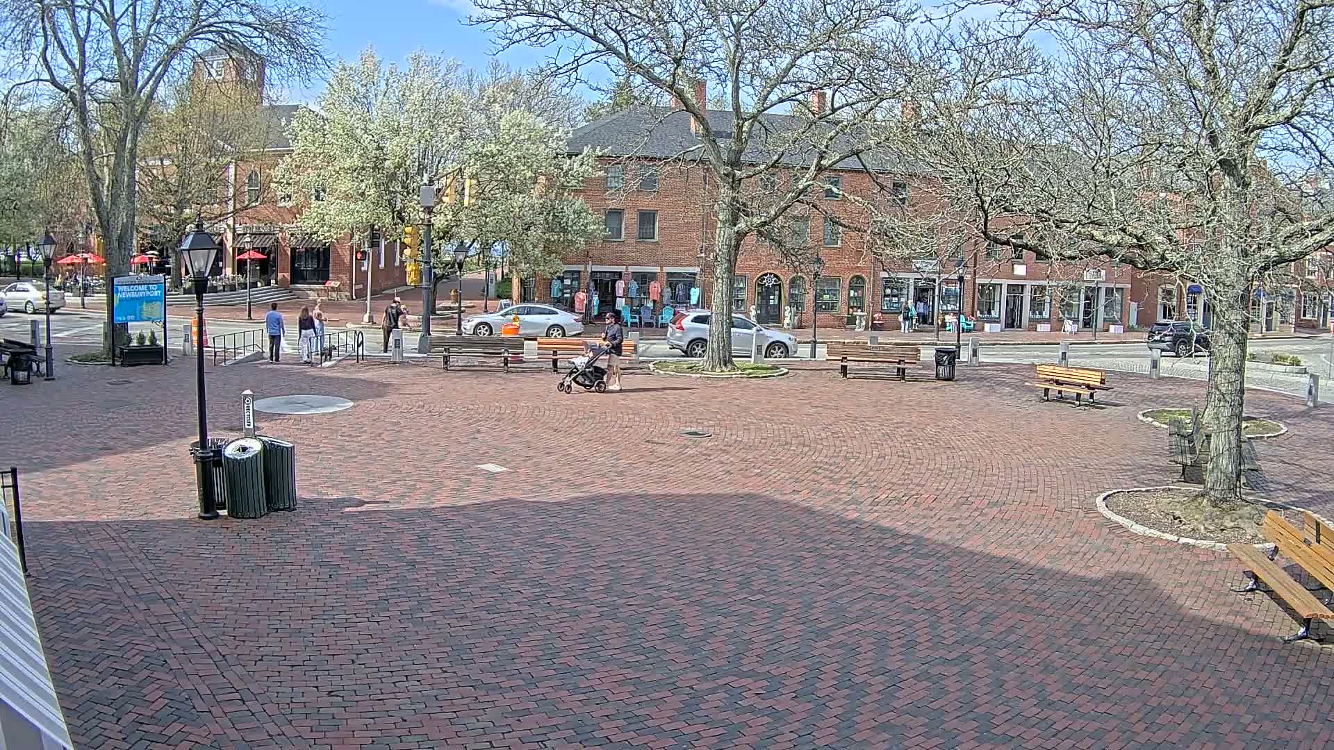 A brick plaza with benches and trees, surrounded by brick buildings under a sunny sky, contains a few pedestrians.