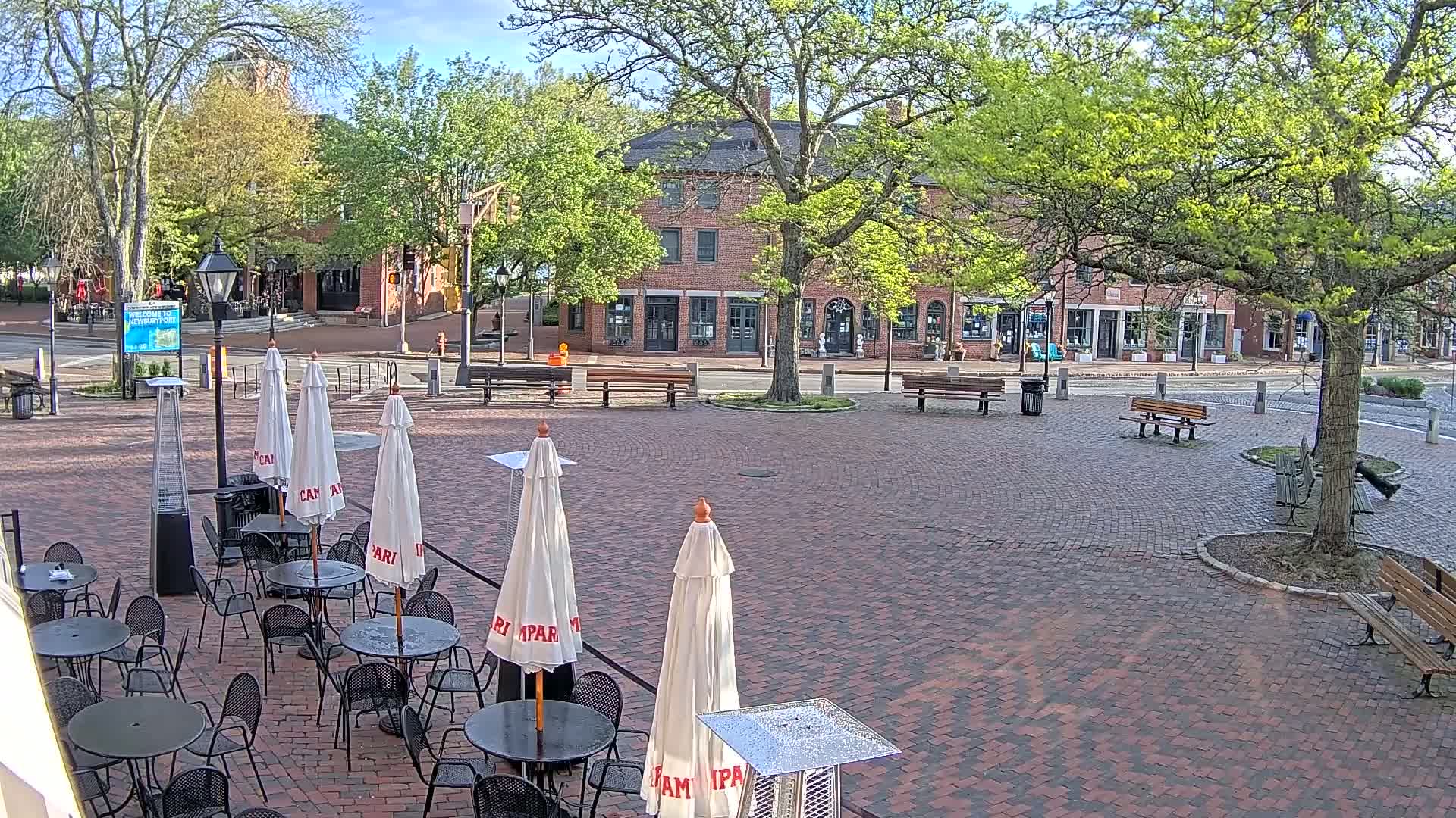 A brick plaza with outdoor seating, patio umbrellas, and park benches sits before a row of brick buildings under a mostly sunny sky.