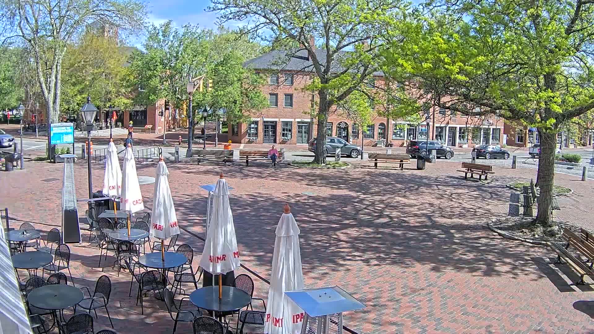 A brick plaza with outdoor seating under white umbrellas, situated in front of brick buildings and leafy trees under a sunny sky.
