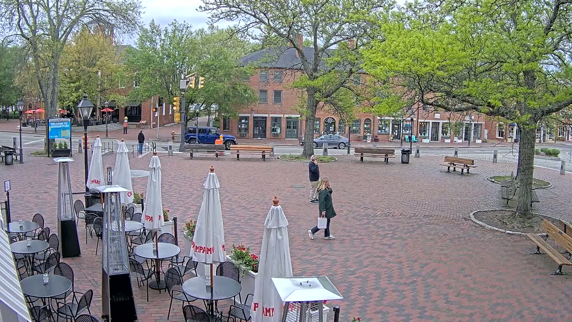 A brick plaza with outdoor café seating, patio heaters, and umbrellas, under an overcast sky, is surrounded by brick buildings and trees.