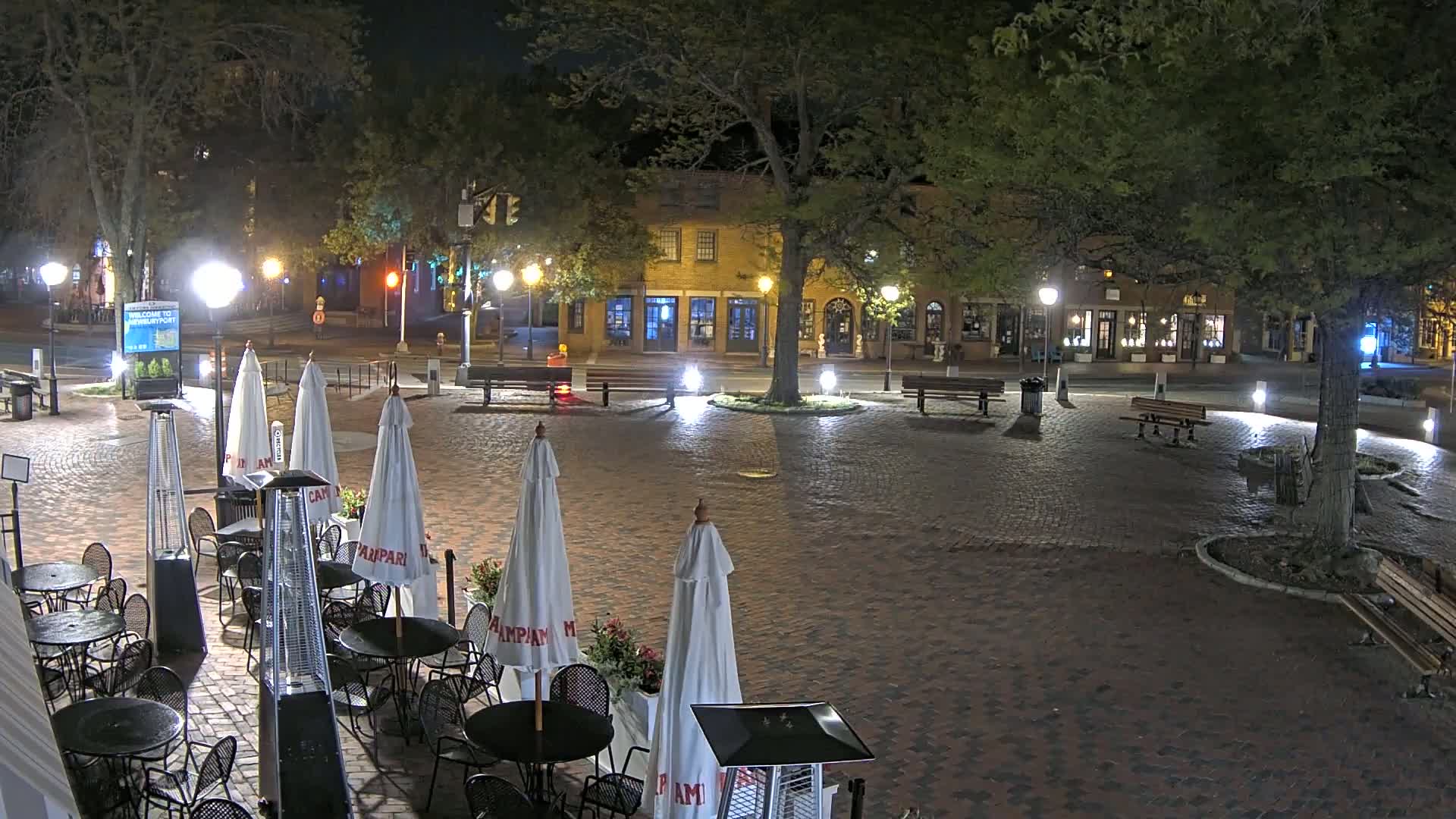 A brick-paved town square at night, illuminated by streetlights, contains outdoor seating with umbrellas and patio heaters near brick buildings under a clear, dark sky.