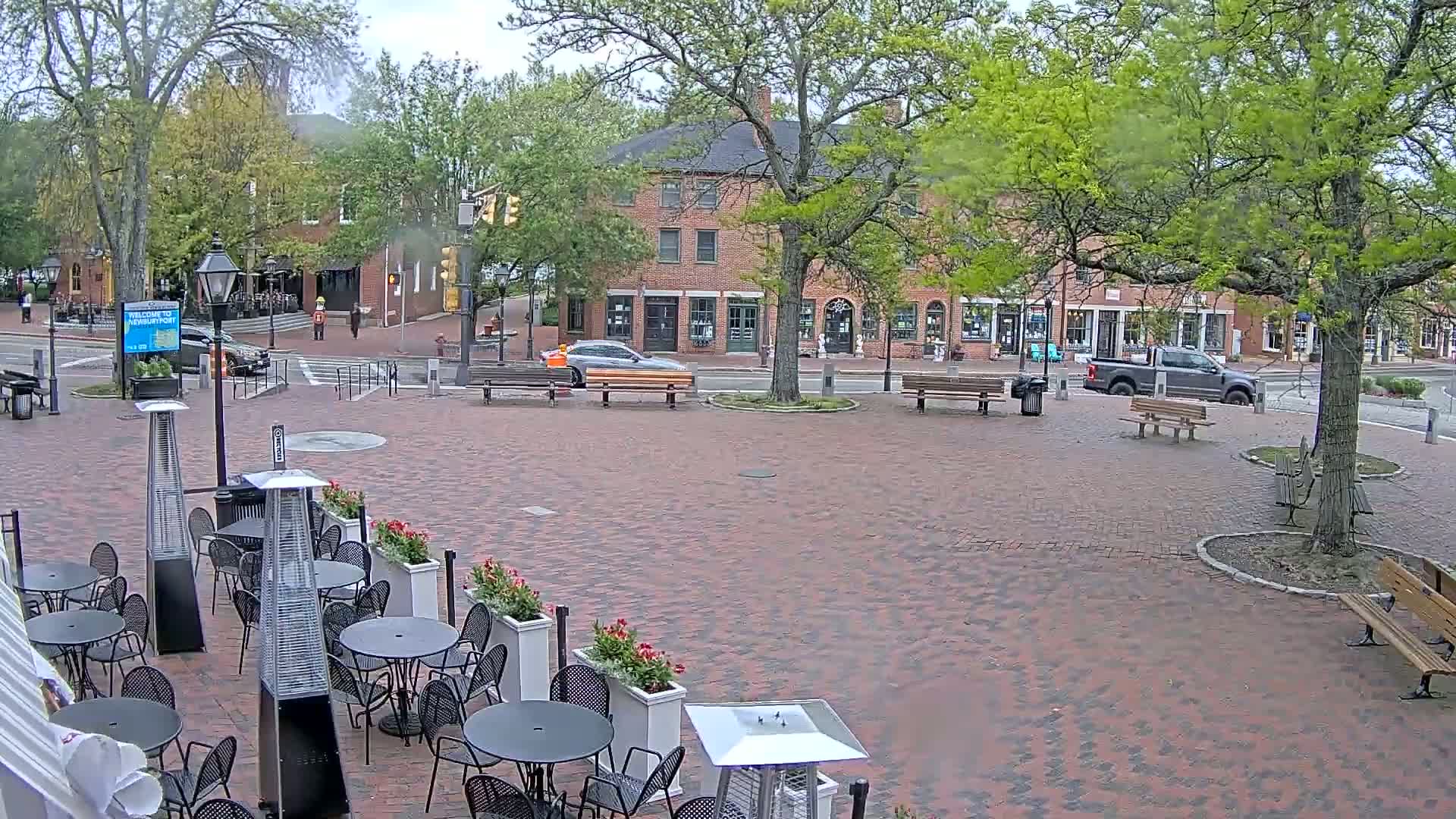 A brick town square with several benches, outdoor seating with patio heaters, and a few cars parked along the street under an overcast sky.