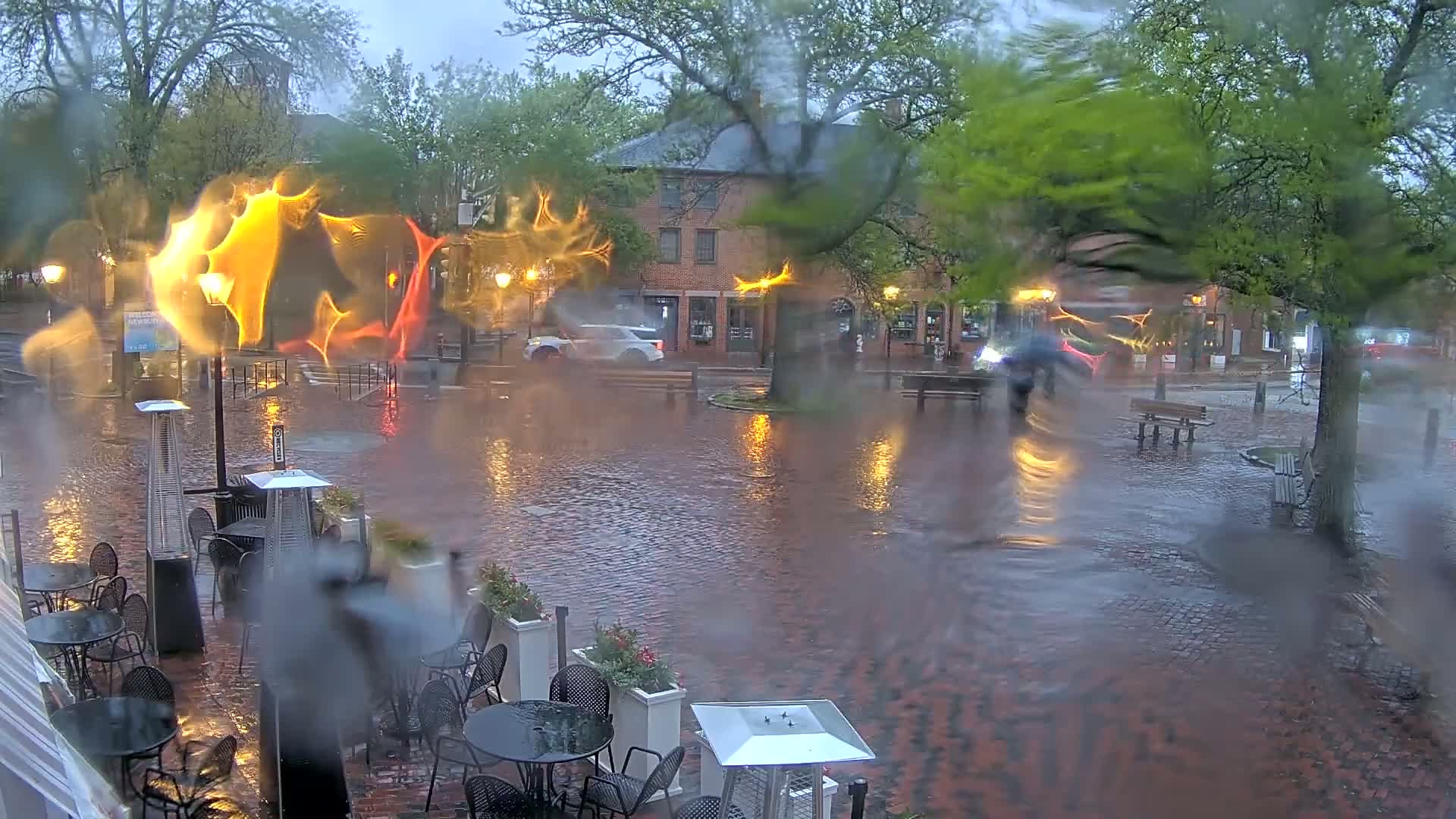 A rain-soaked brick plaza with outdoor patio furniture and street lights is viewed through a rain-covered window, showing cars driving past brick buildings under a gray sky.