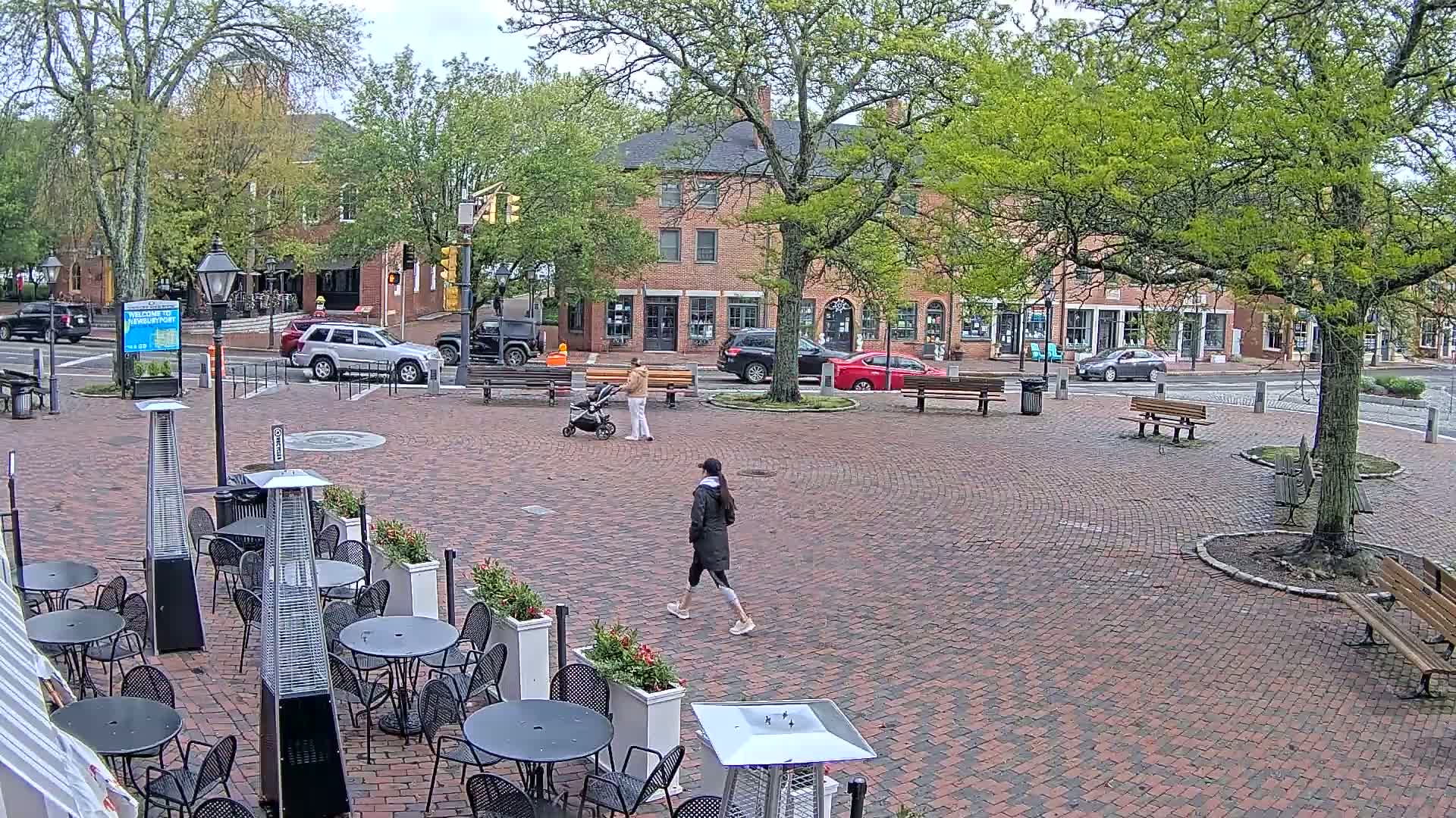 A brick town square with benches, trees, and outdoor patio furniture, under an overcast sky.