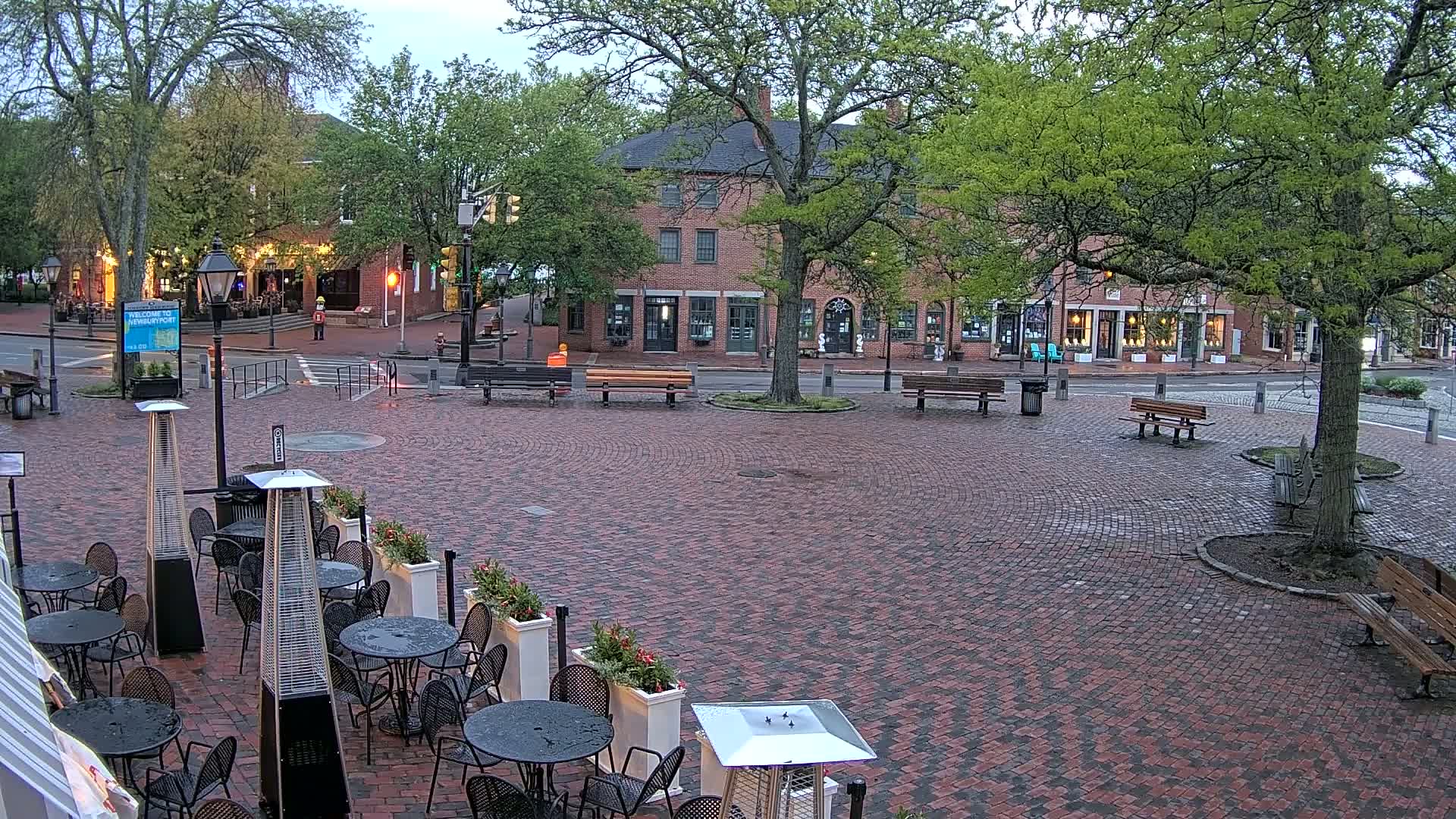 A brick town square with several benches and outdoor restaurant seating, under an overcast sky.