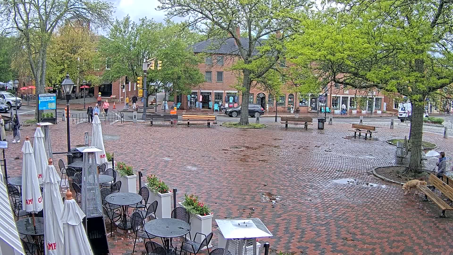 A brick town square with outdoor seating, trees, and shops under a cloudy sky, with some puddles on the ground.