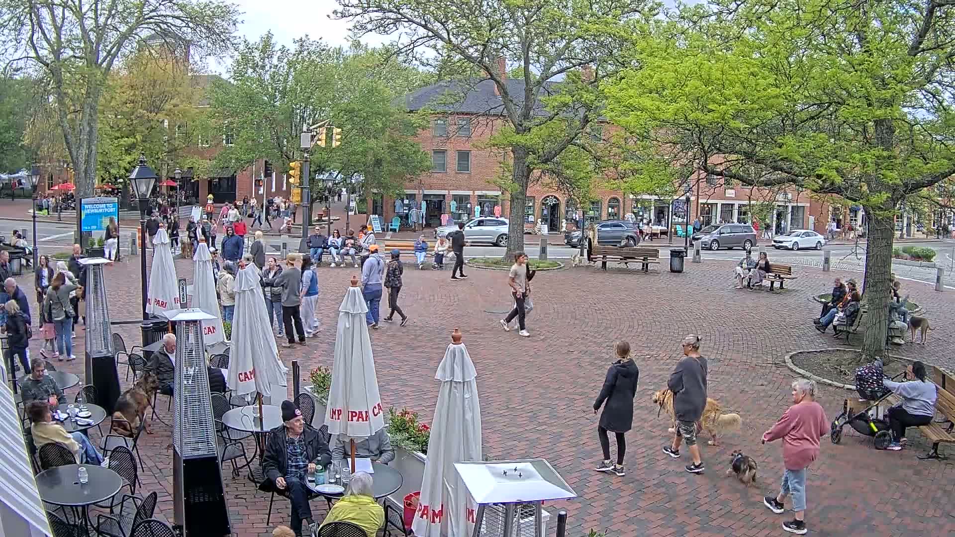 A brick town square with people walking, sitting on benches, and eating at outdoor cafes under partly cloudy skies.