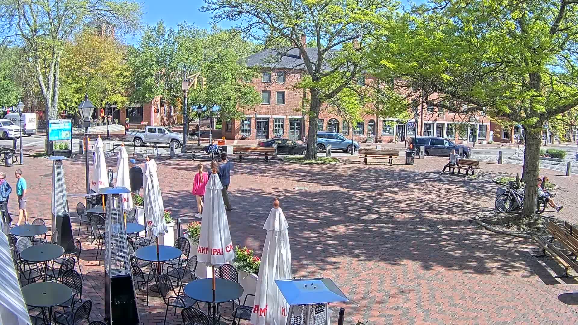 A sunny outdoor brick plaza with several people walking and sitting, surrounded by trees and brick buildings, with patio furniture including umbrellas and patio heaters.