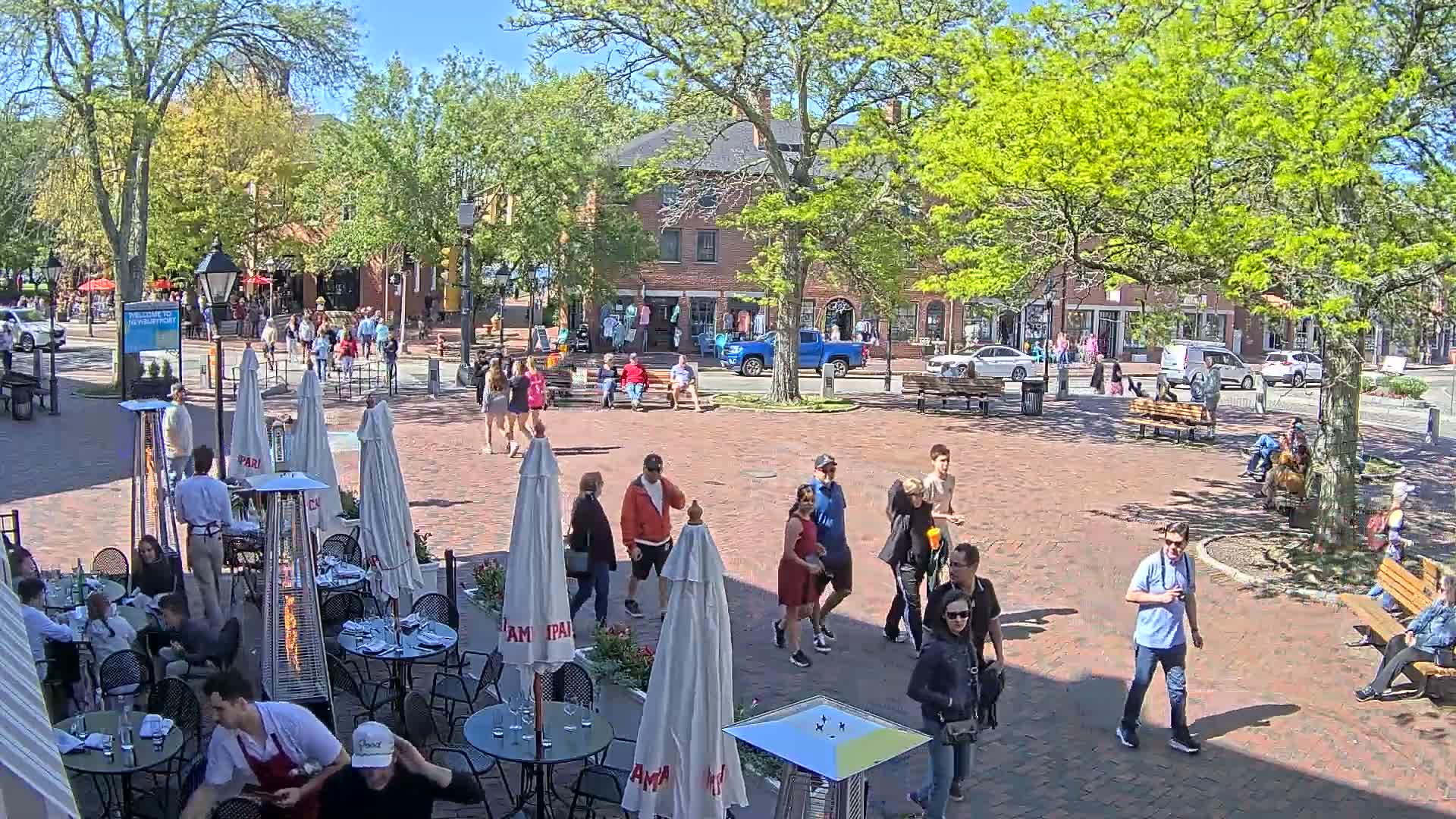 A brick town square, under a sunny sky, is filled with people walking and sitting at outdoor cafe tables.