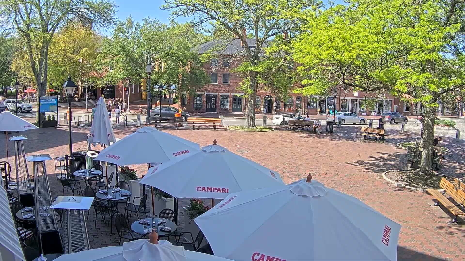 A brick plaza with outdoor cafe seating under white umbrellas, surrounded by brick buildings and leafy green trees on a sunny day.