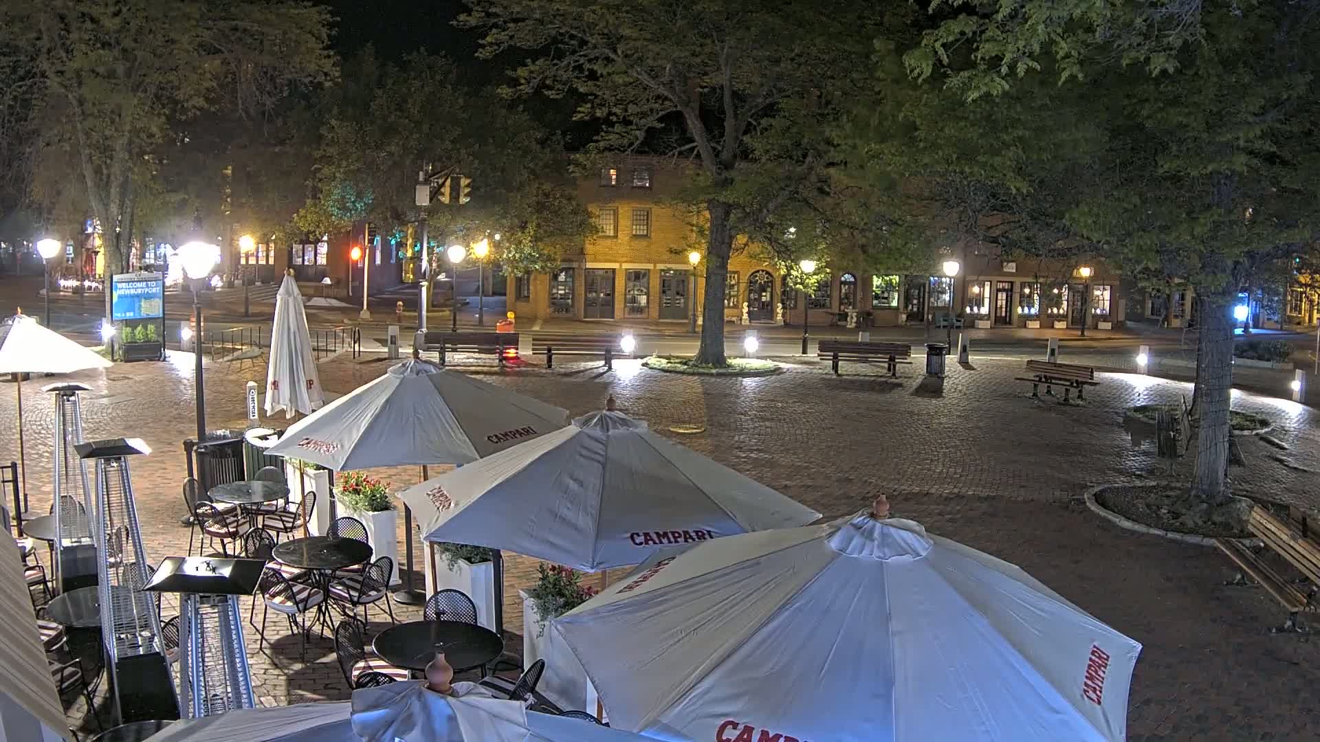A brick plaza at night, lit by streetlamps, contains several outdoor cafe umbrellas and patio heaters, with shops and trees visible in the background.