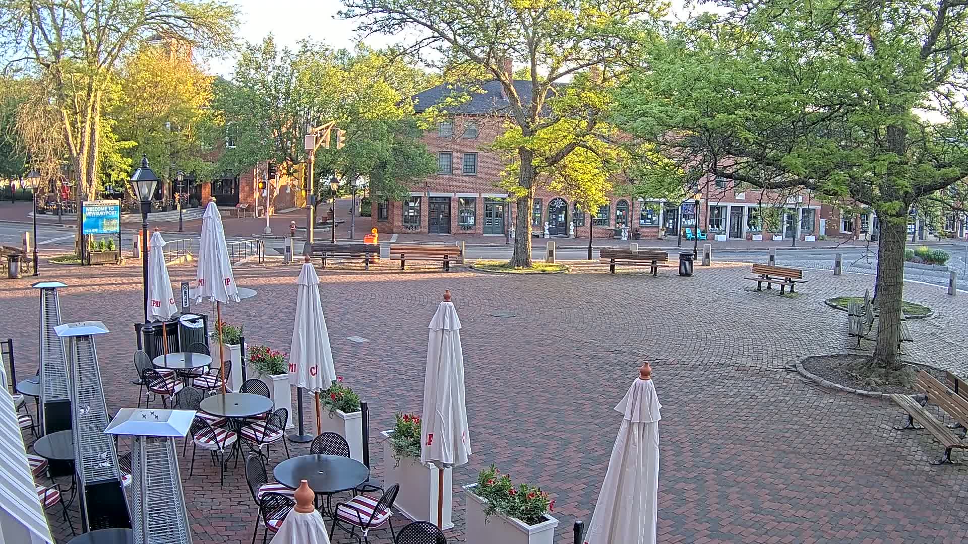 A brick paved town square with outdoor cafe seating, white umbrellas, and patio heaters, under a mostly sunny sky with lush green trees and brick buildings in the background.