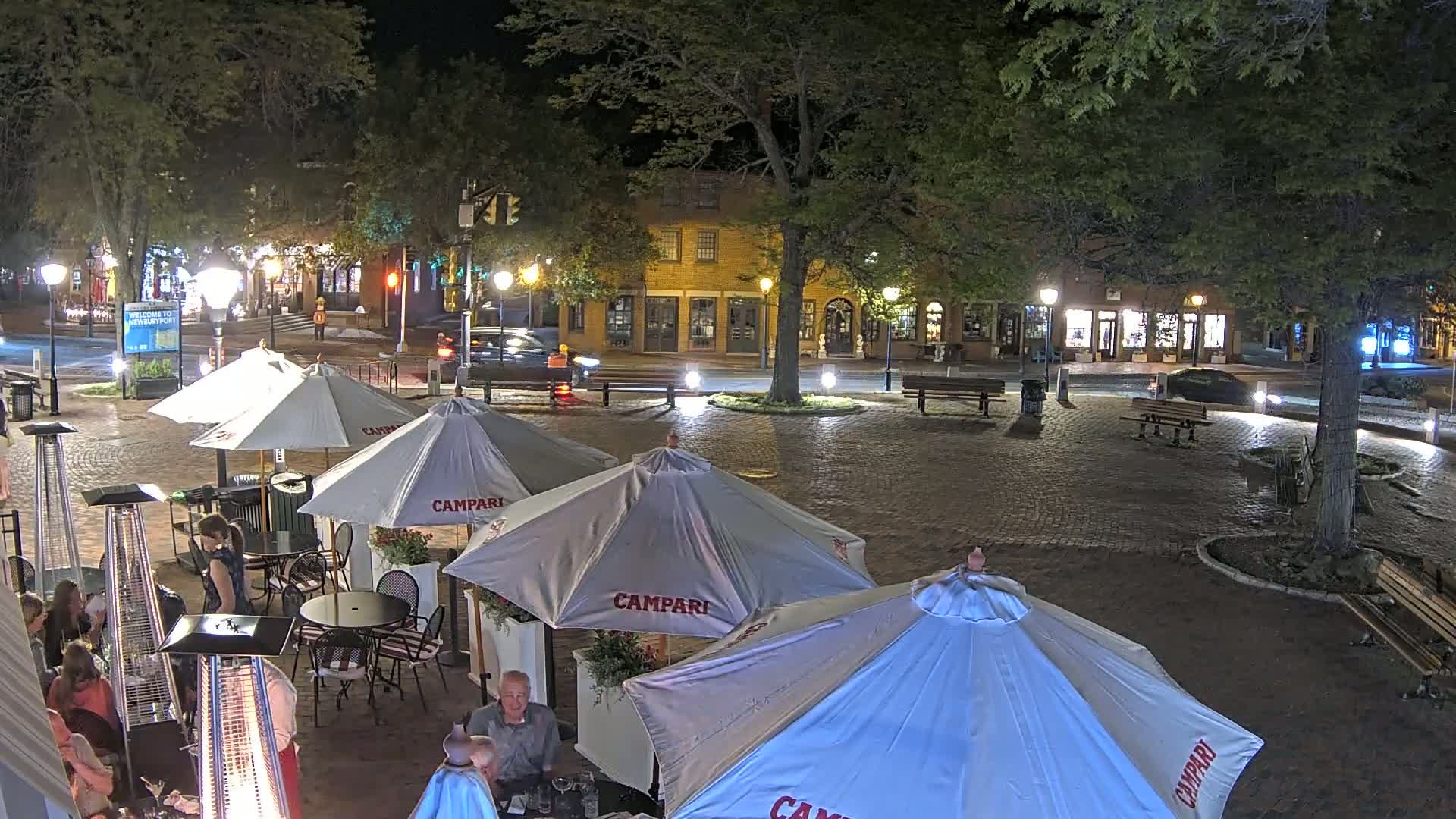 A nighttime outdoor scene of a brick plaza with several patio umbrellas, heat lamps, and tables set up around a central tree, adjacent to buildings with street lights illuminating the area.