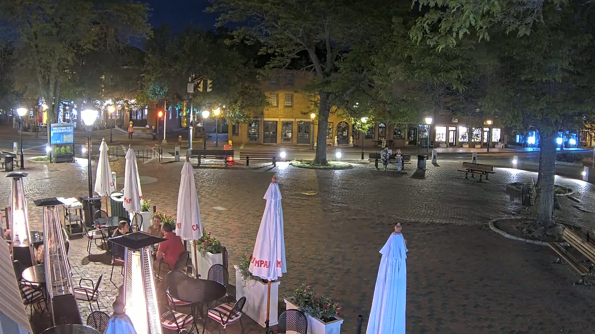 A nighttime outdoor scene shows a brick plaza with cafe seating, patio umbrellas, and heat lamps, surrounded by buildings and trees under a clear sky.