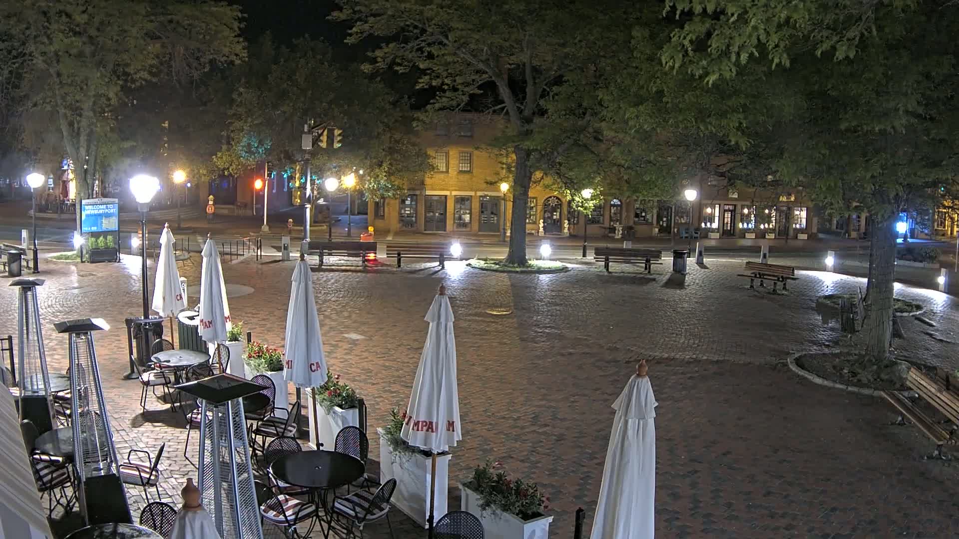 A brick plaza at night, illuminated by streetlights, contains several park benches, outdoor cafe seating with patio heaters and umbrellas, and trees.