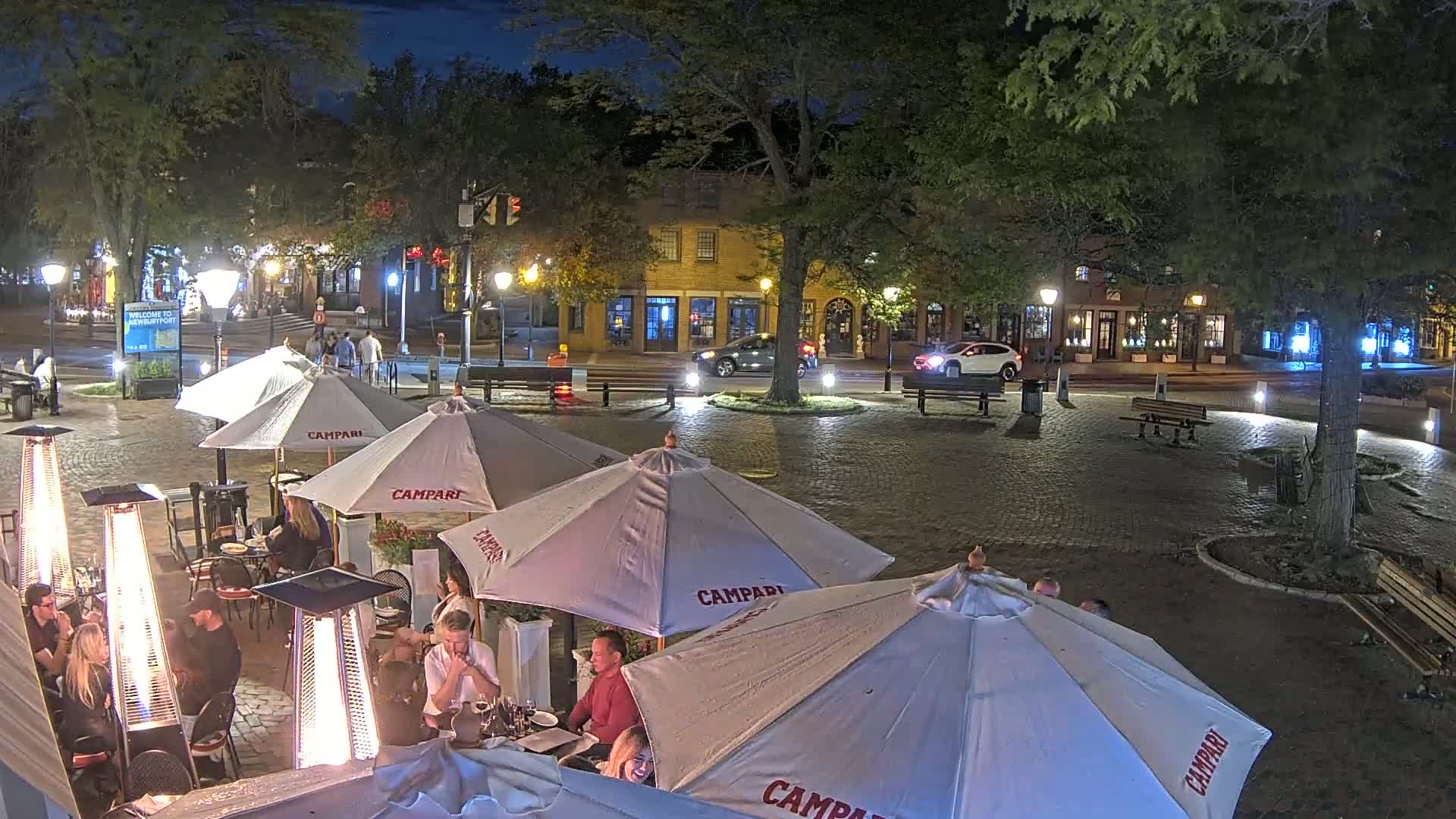 A nighttime outdoor patio scene with several tables under umbrellas, lit by patio heaters and streetlights, situated in a cobblestone town square near buildings.