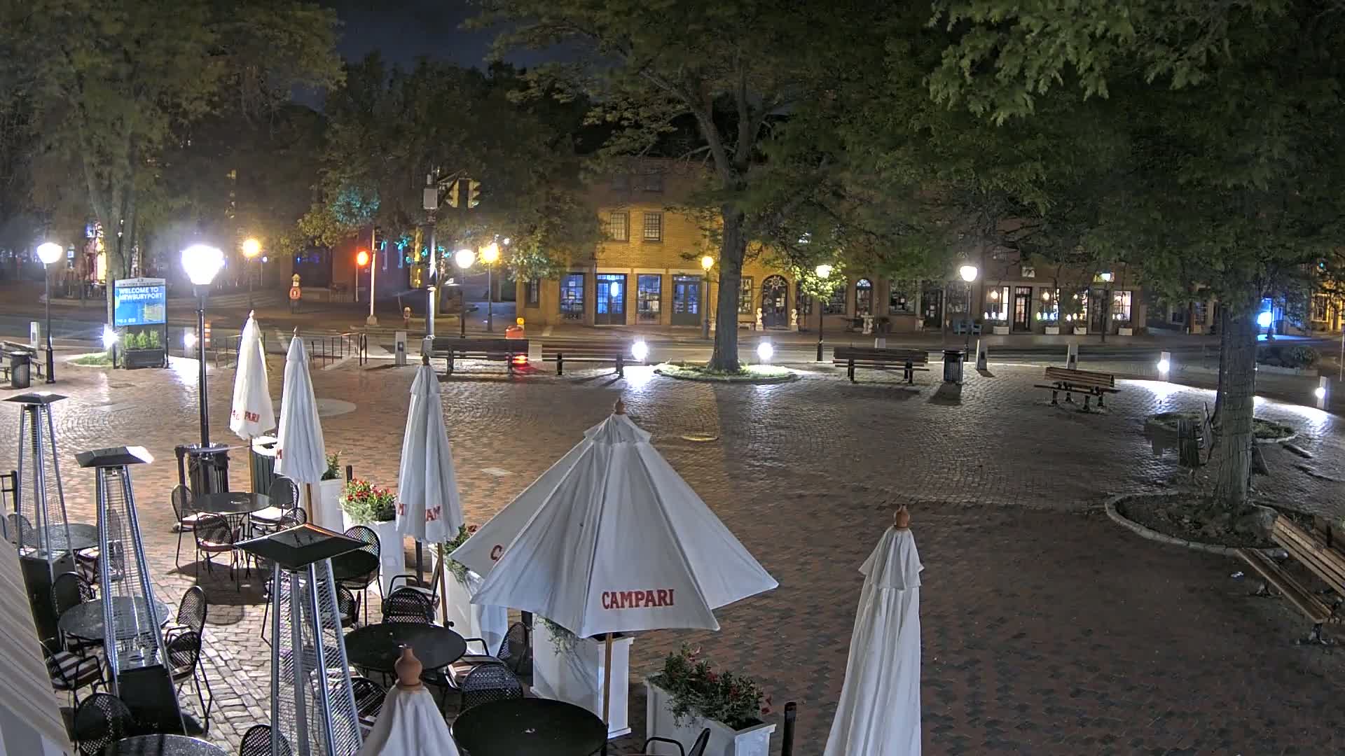 A brick-paved outdoor area at night, illuminated by streetlights, features several cafe umbrellas, patio heaters, and tables, with buildings and trees in the background under a clear, dark sky.