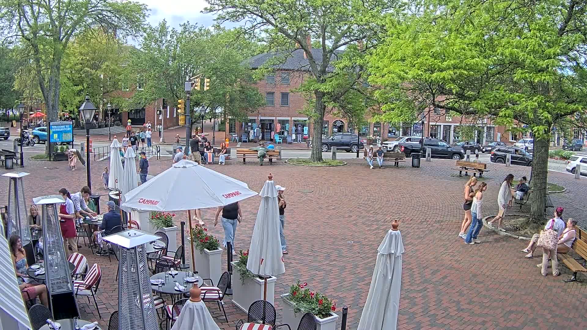 A brick town square with outdoor seating at restaurants, people walking around, and parked cars lining the streets under a partly sunny sky.