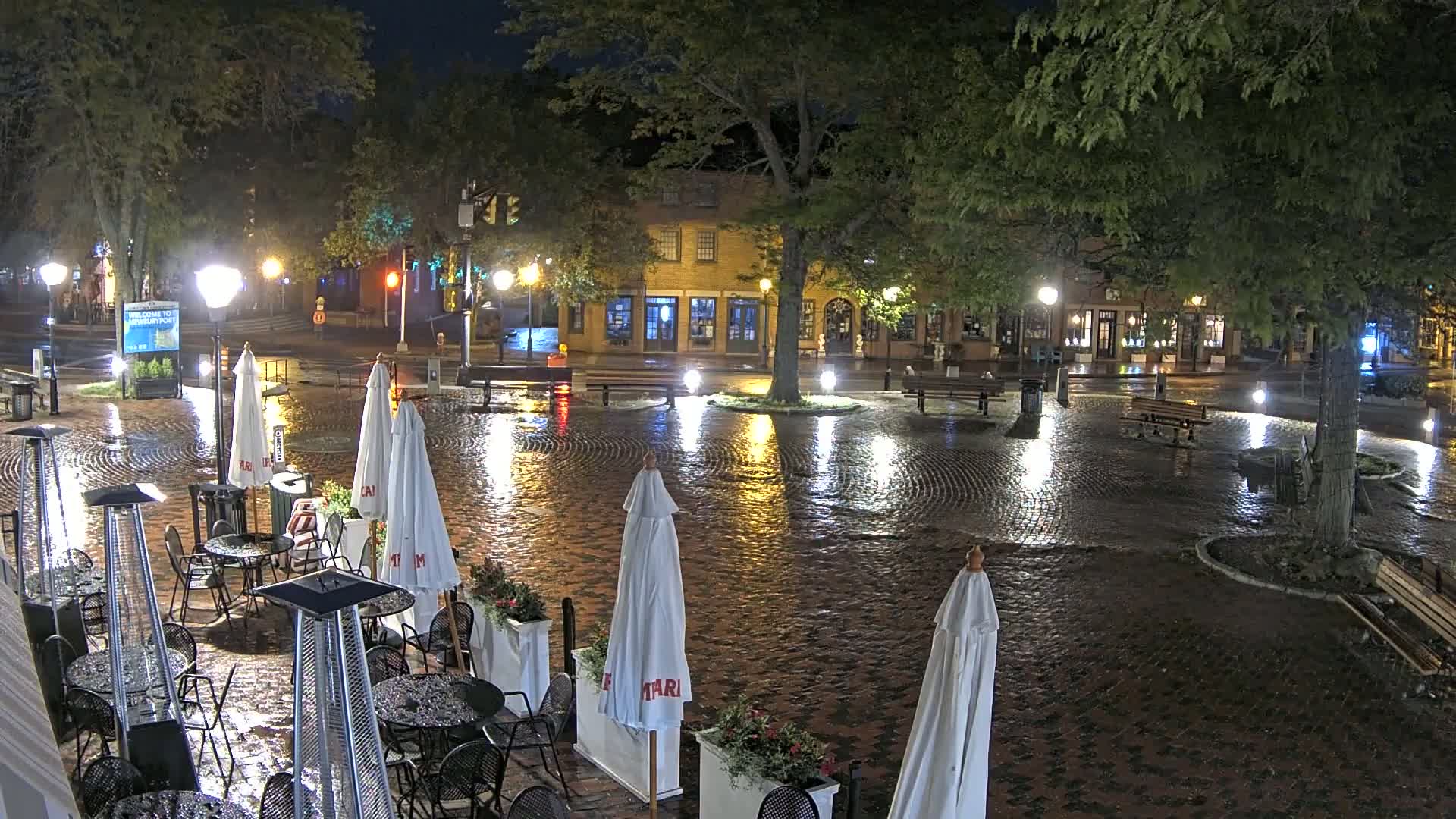 A rain-slicked brick town square at night, illuminated by streetlights, features outdoor seating areas with umbrellas and patio heaters near buildings.