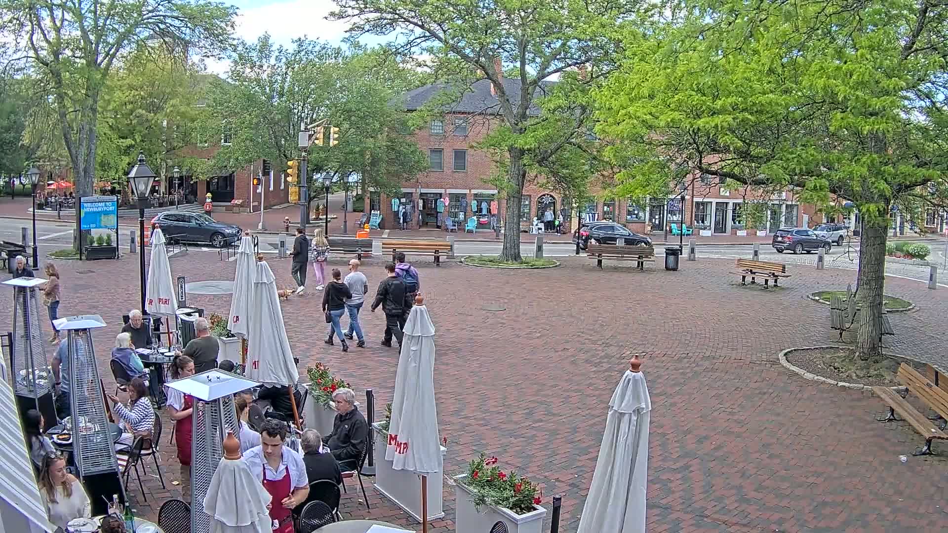 A brick plaza with outdoor seating and several people walking by, under a partly cloudy sky.