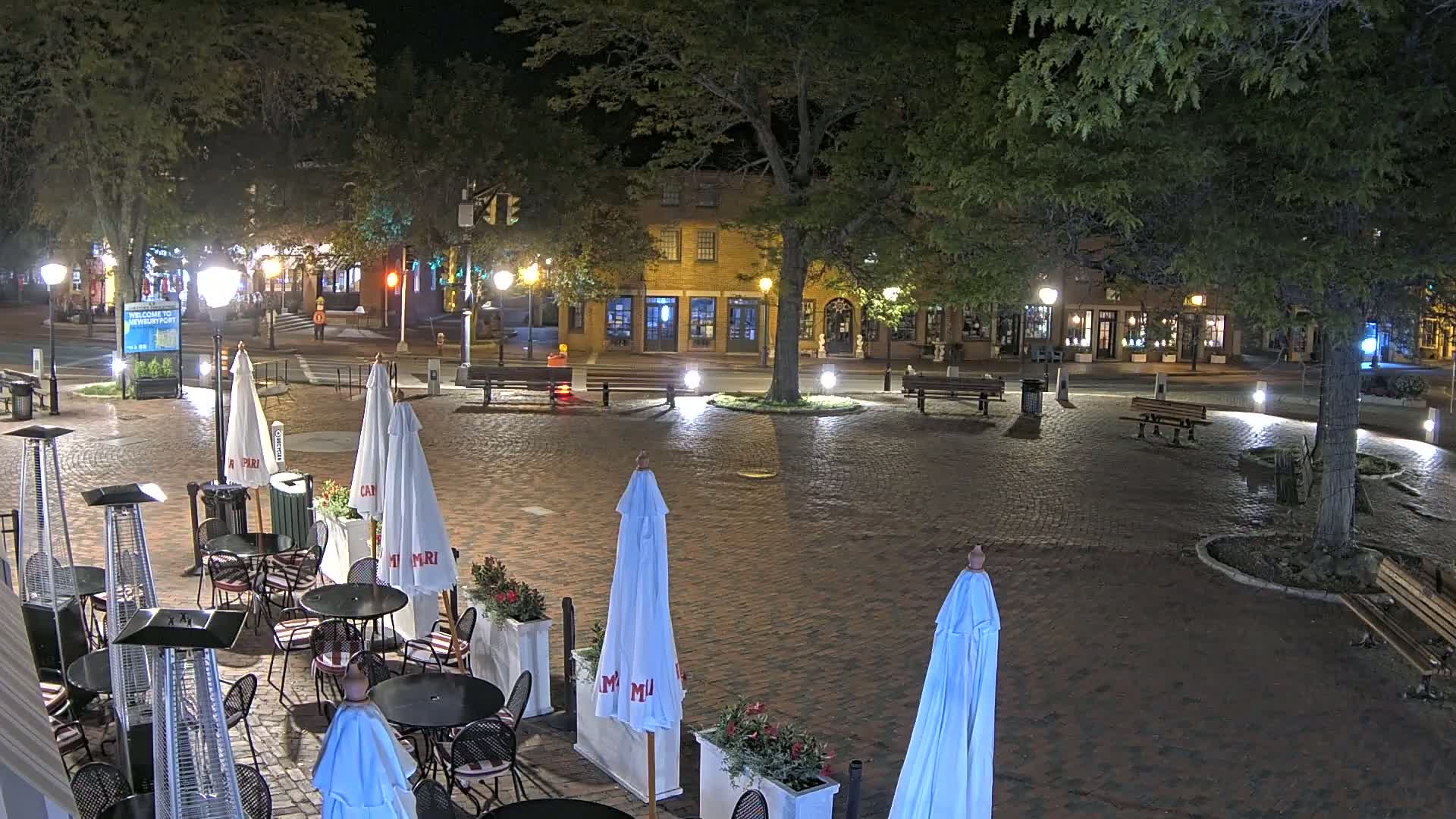 A brick town square at night, illuminated by streetlights, contains outdoor cafe seating with umbrellas and patio heaters, surrounded by trees and buildings.