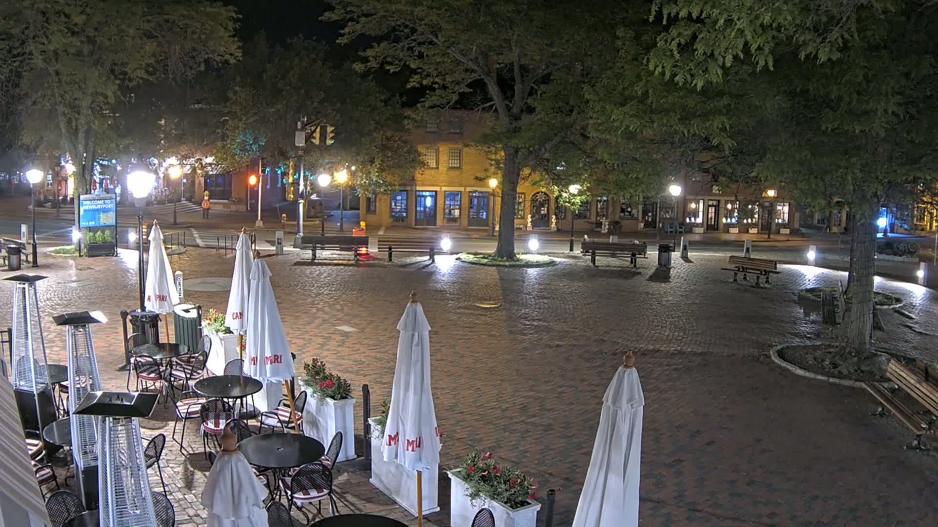 A brick town square at night, illuminated by streetlights, features several outdoor café tables with umbrellas and patio heaters under the shade of large trees.