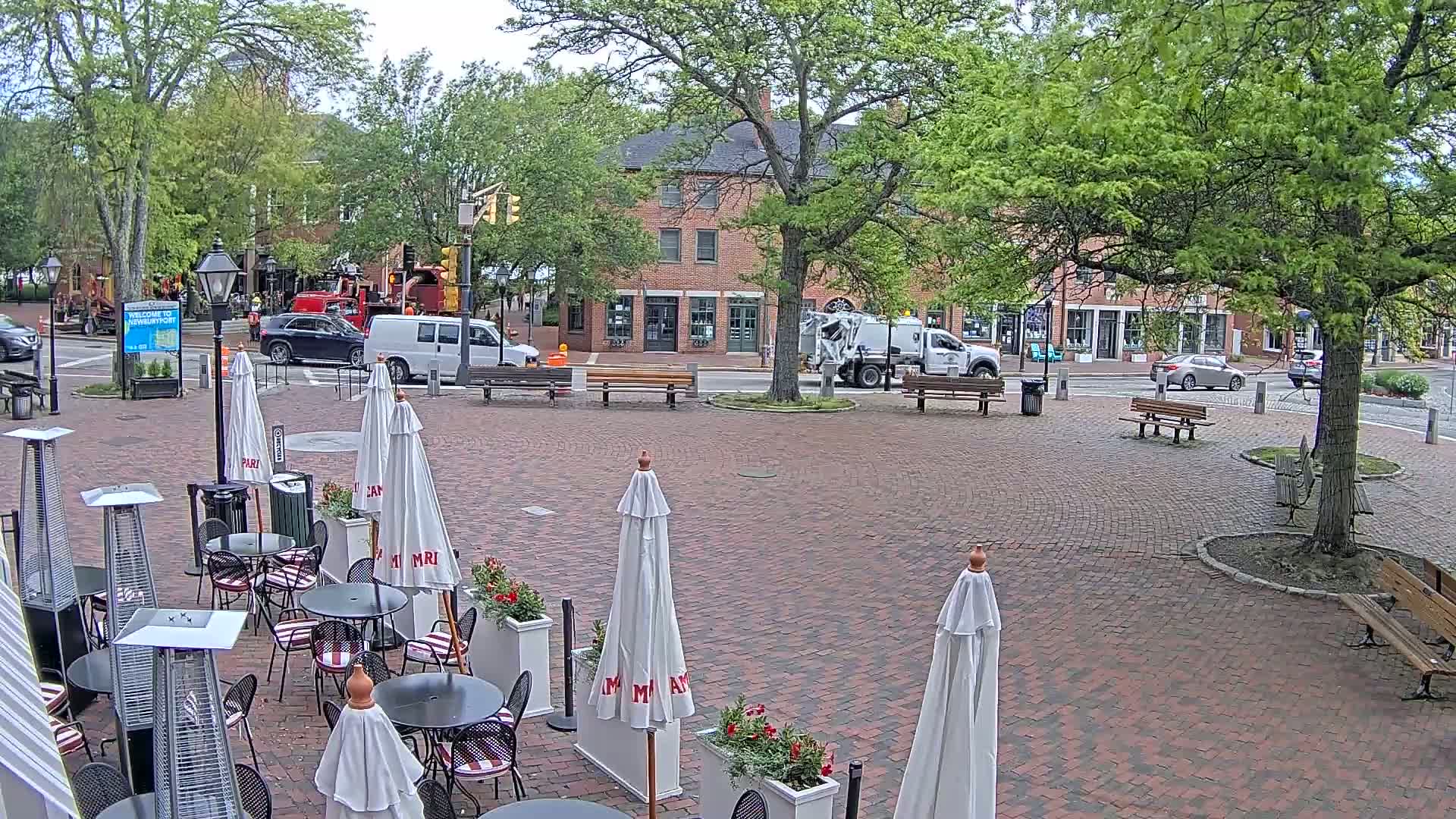 A brick plaza with outdoor cafe seating under umbrellas, surrounded by brick buildings and lush green trees on a partly cloudy day.