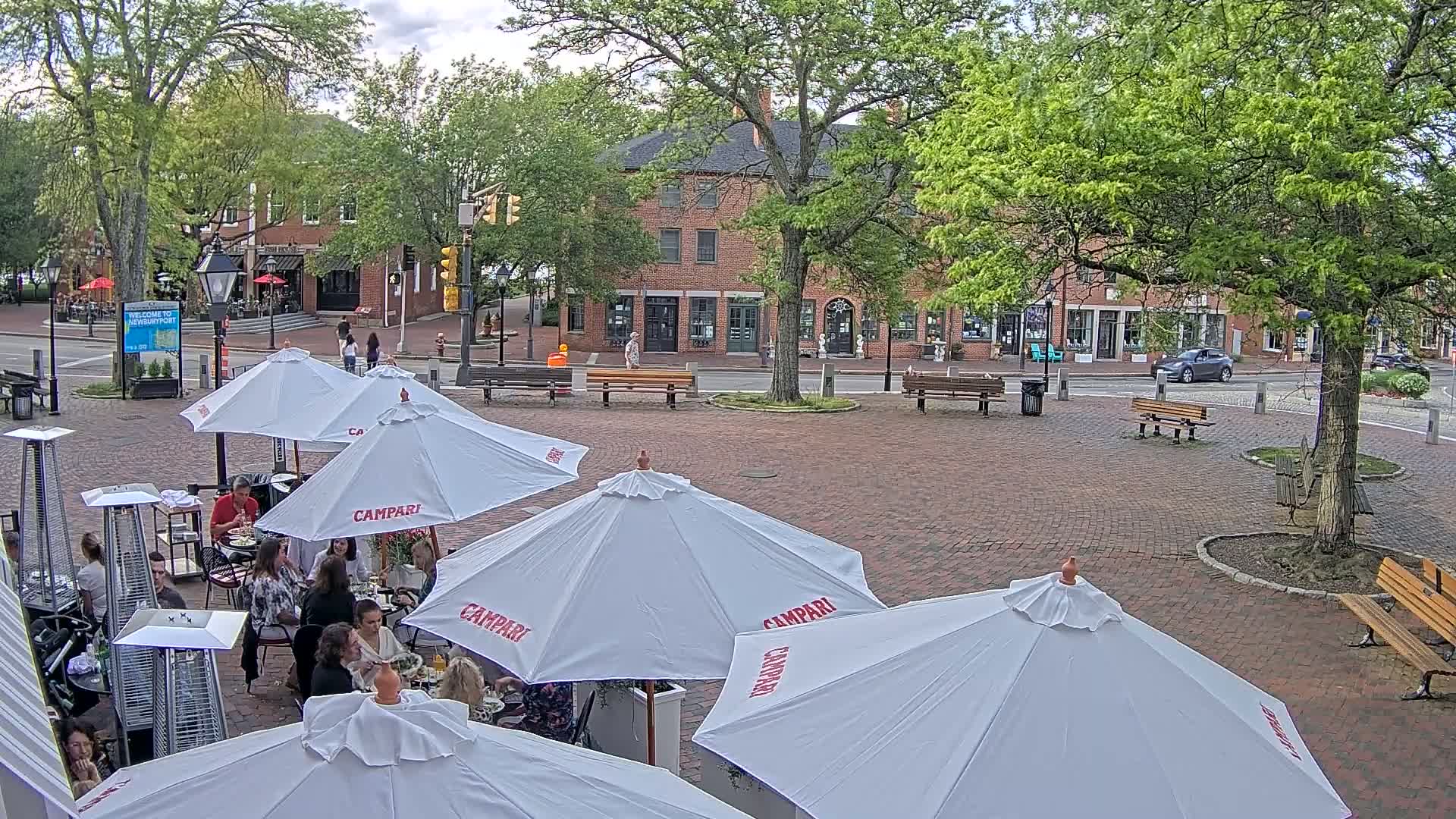 An outdoor brick patio with several white umbrellas shading cafe tables and patrons, surrounded by trees and brick buildings on a partly cloudy day.