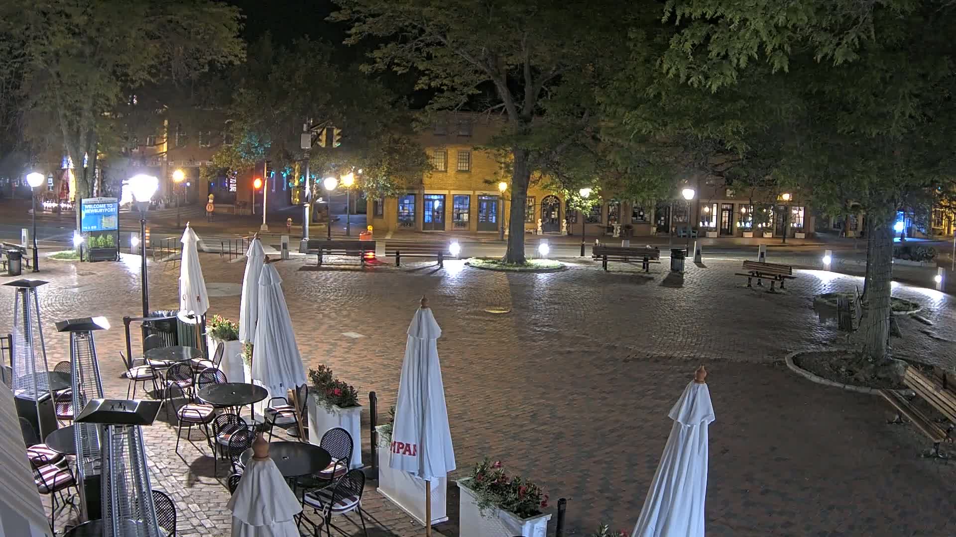 A brick plaza at night, illuminated by streetlights, contains outdoor cafe seating with umbrellas and patio heaters, and is surrounded by buildings and trees.