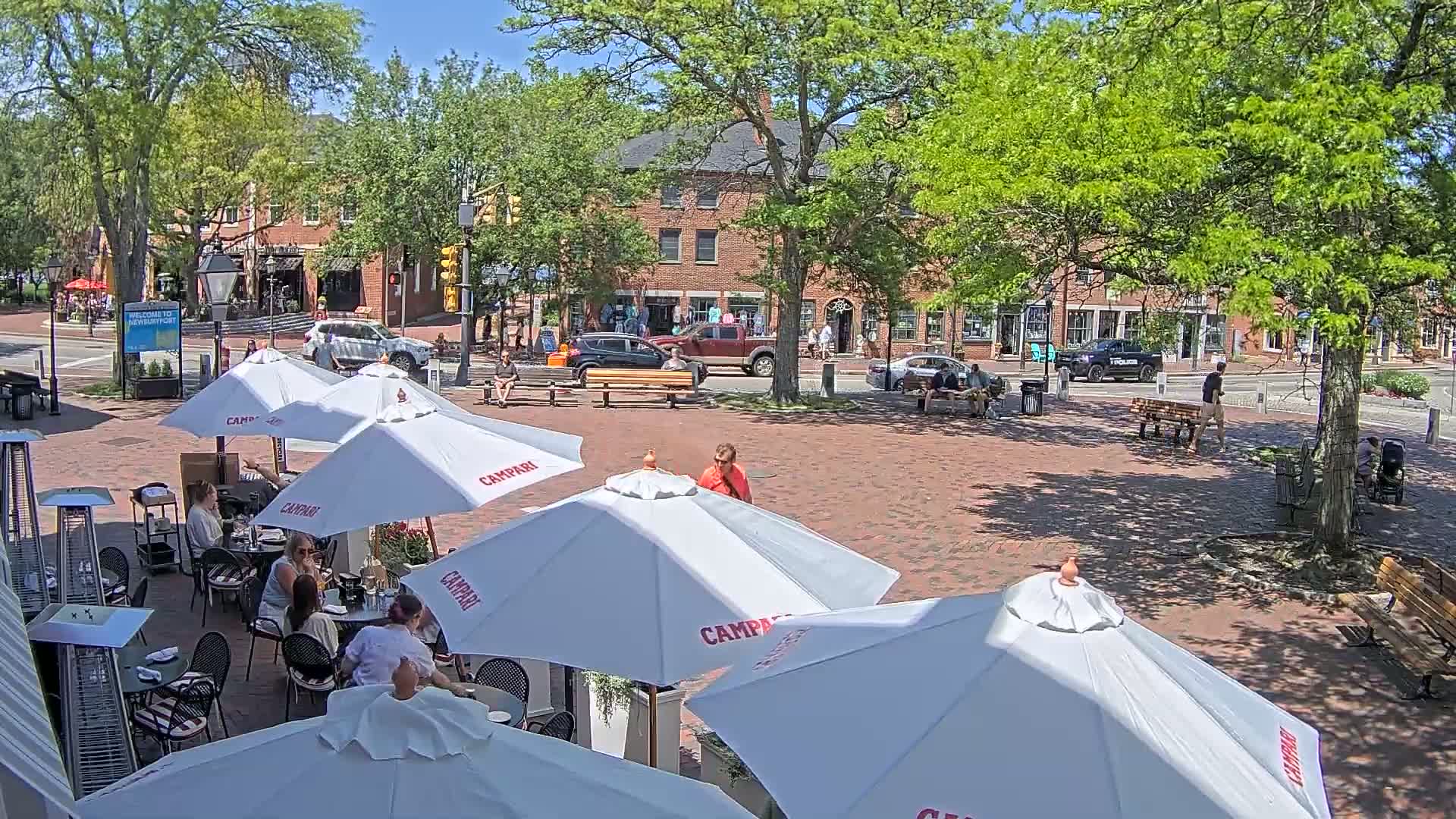 A sunny outdoor cafe with white umbrellas, patrons seated at tables, and brick paving, situated in front of brick buildings lined with trees.