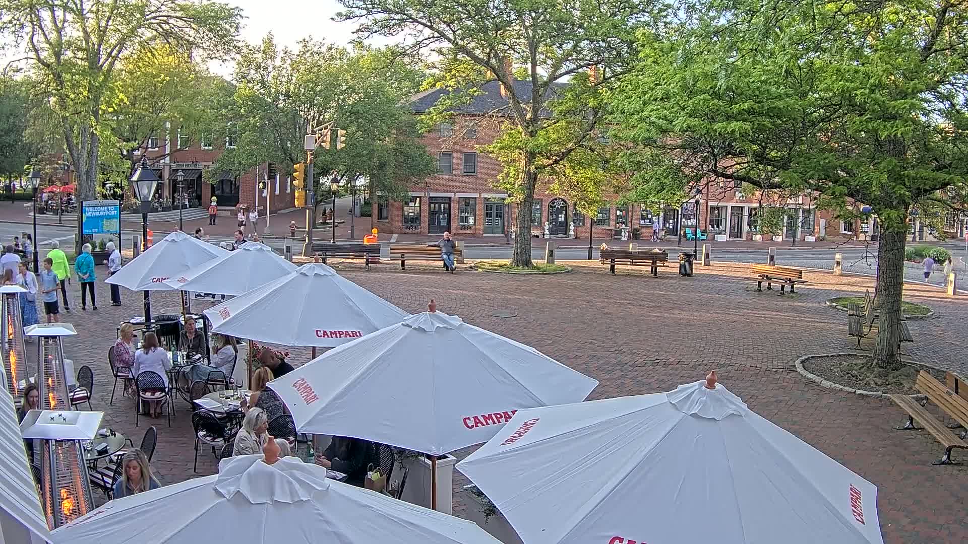 An outdoor patio area with white umbrellas and patio heaters, surrounded by brick pavement, park benches, and leafy green trees, in front of a brick building on a partly sunny day.