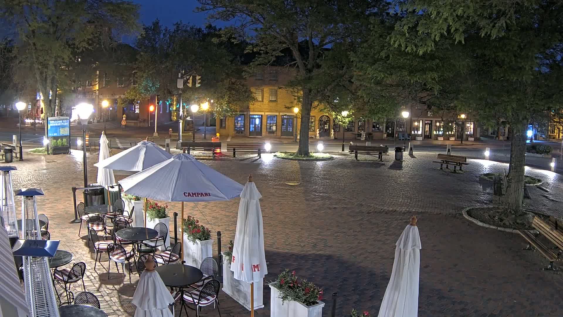 A brick plaza at night, illuminated by streetlights, contains several outdoor cafe tables and umbrellas, with brick buildings and trees in the background.