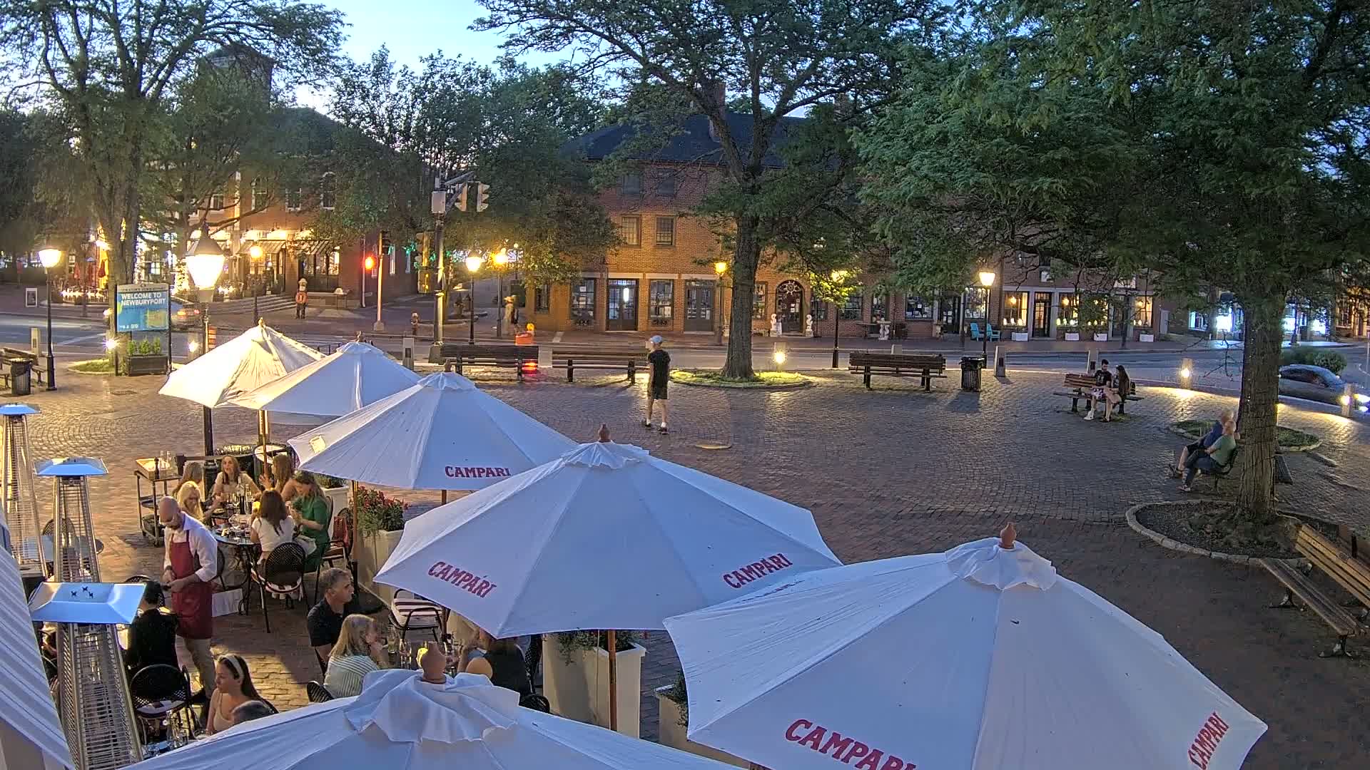 An outdoor brick plaza at dusk, with several groups of people seated at tables under white umbrellas at a restaurant, surrounded by buildings and trees.