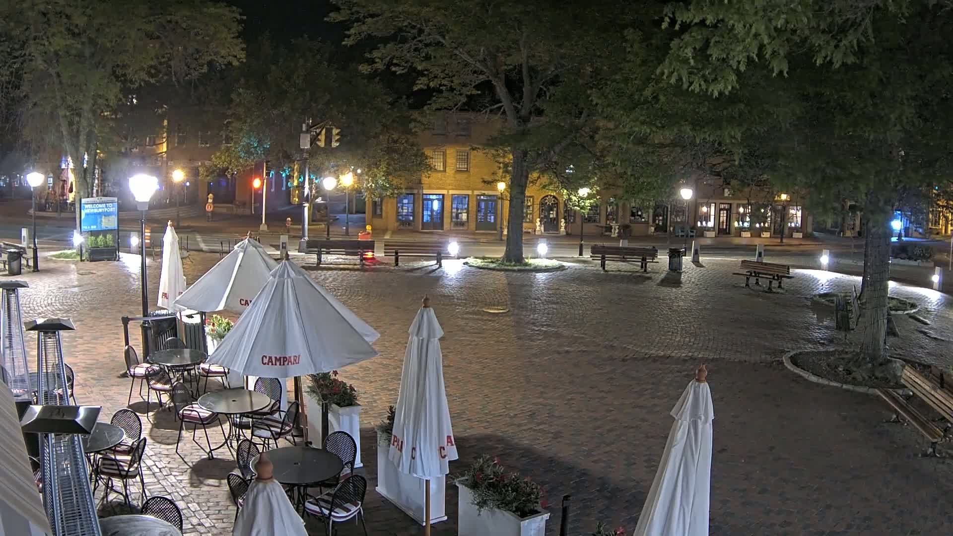 A brick-paved town square at night, illuminated by streetlights, features outdoor cafe seating with umbrellas, benches, and trees.