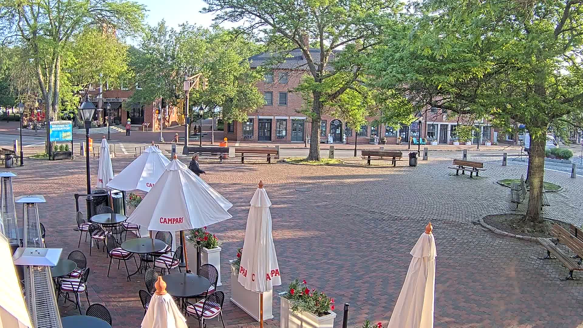 A brick plaza with outdoor cafe seating under white umbrellas, surrounded by trees and a brick building in the background on a sunny day.