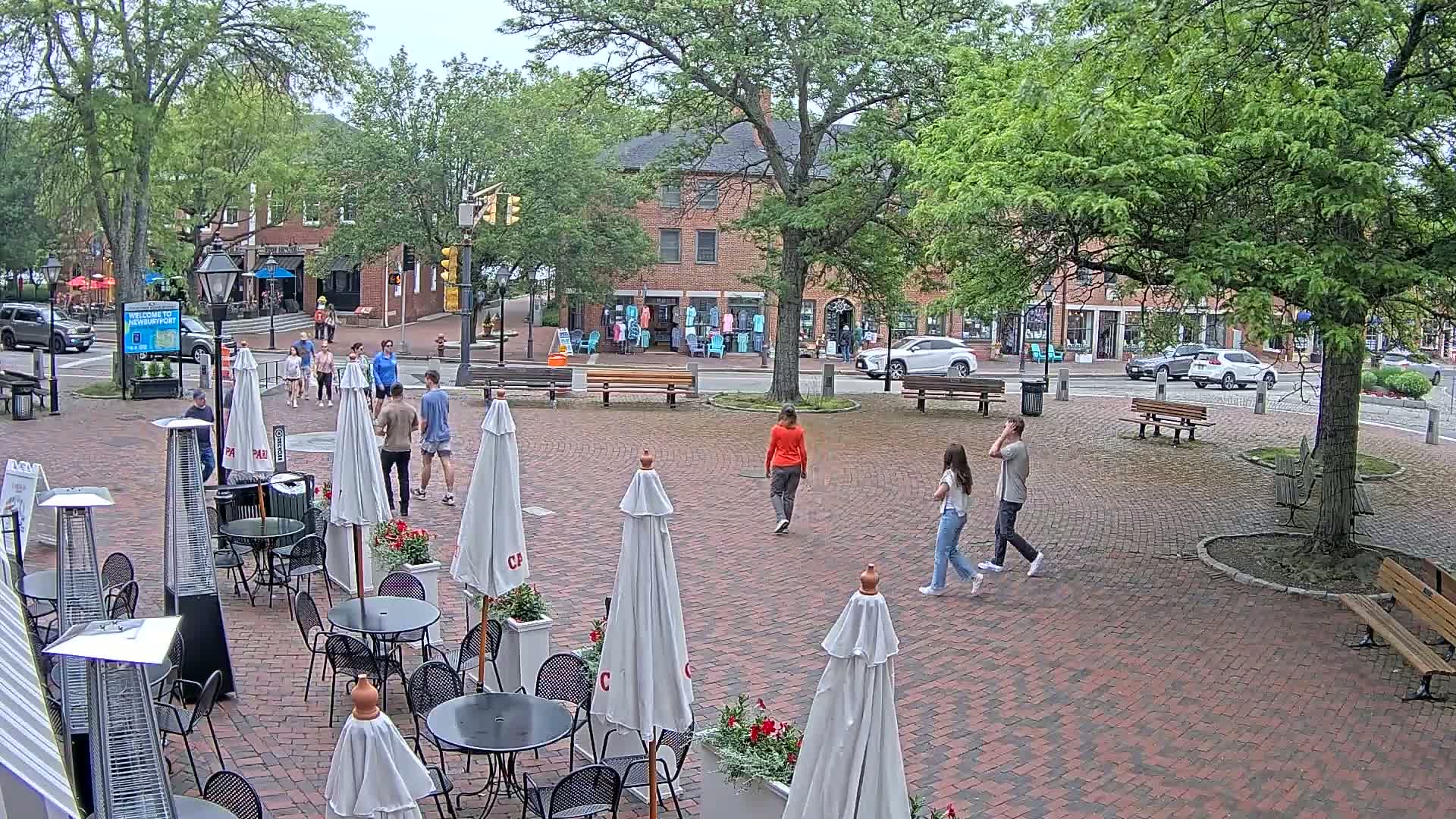 A brick plaza with outdoor seating, several pedestrians, and brick buildings in the background under a partly cloudy sky.