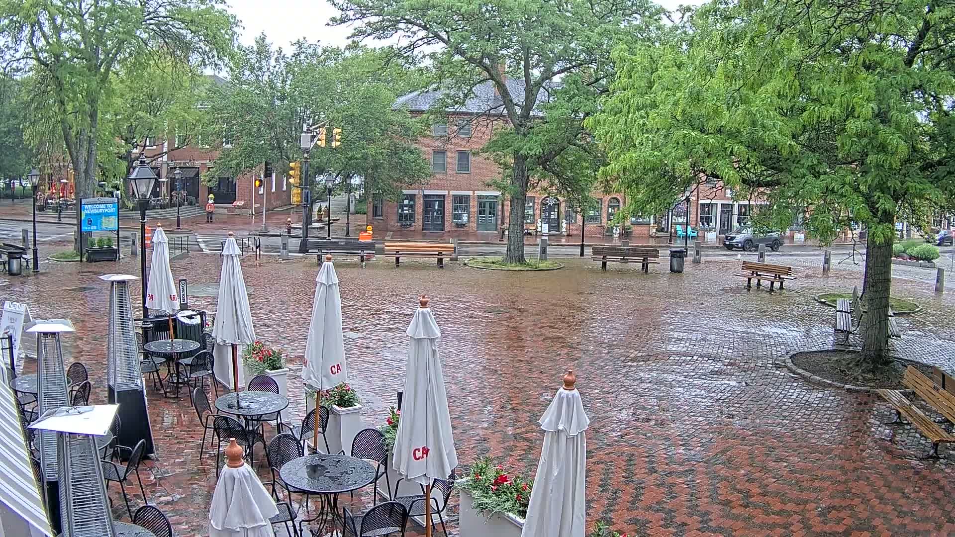 A rain-slicked brick plaza with outdoor cafe seating under umbrellas, surrounded by trees and brick buildings.