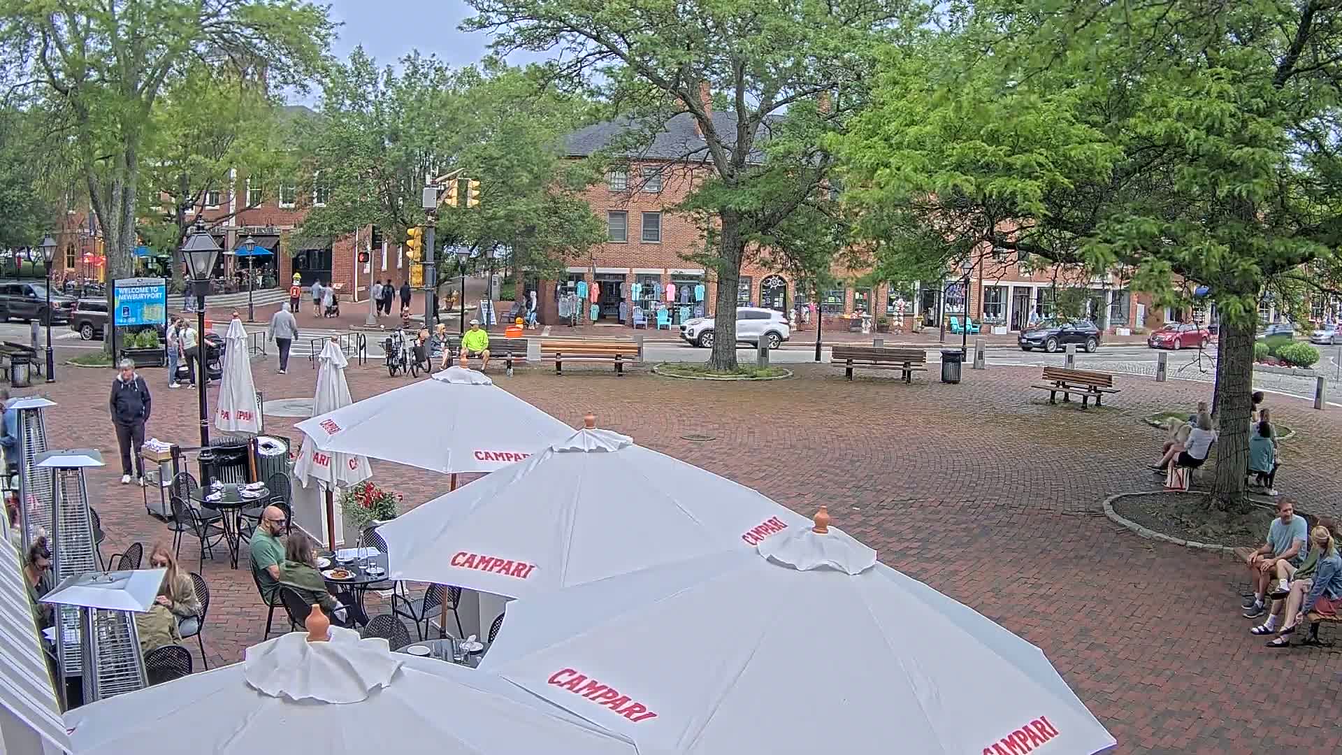 An outdoor brick plaza with several people sitting at cafe tables under umbrellas and others walking around, all under an overcast sky.