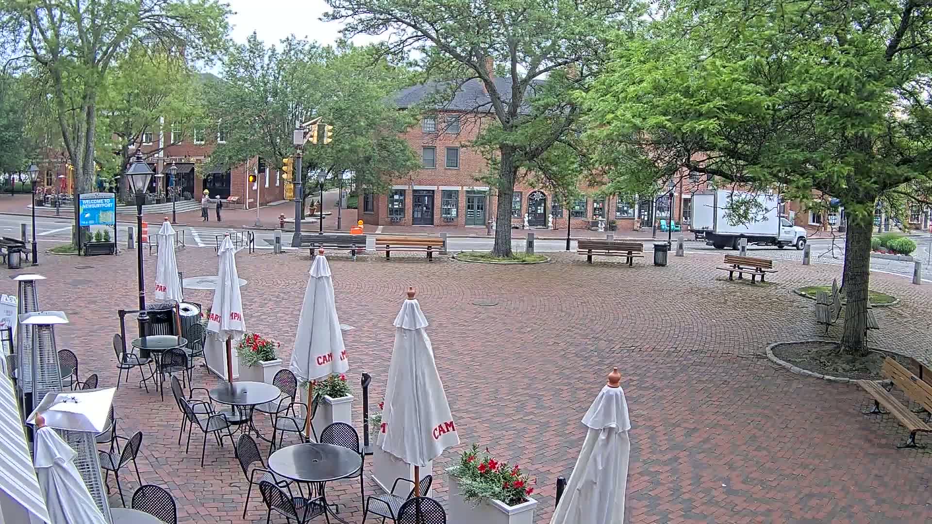 A brick plaza with outdoor cafe seating, several trees, and a brick building in the background on an overcast day.