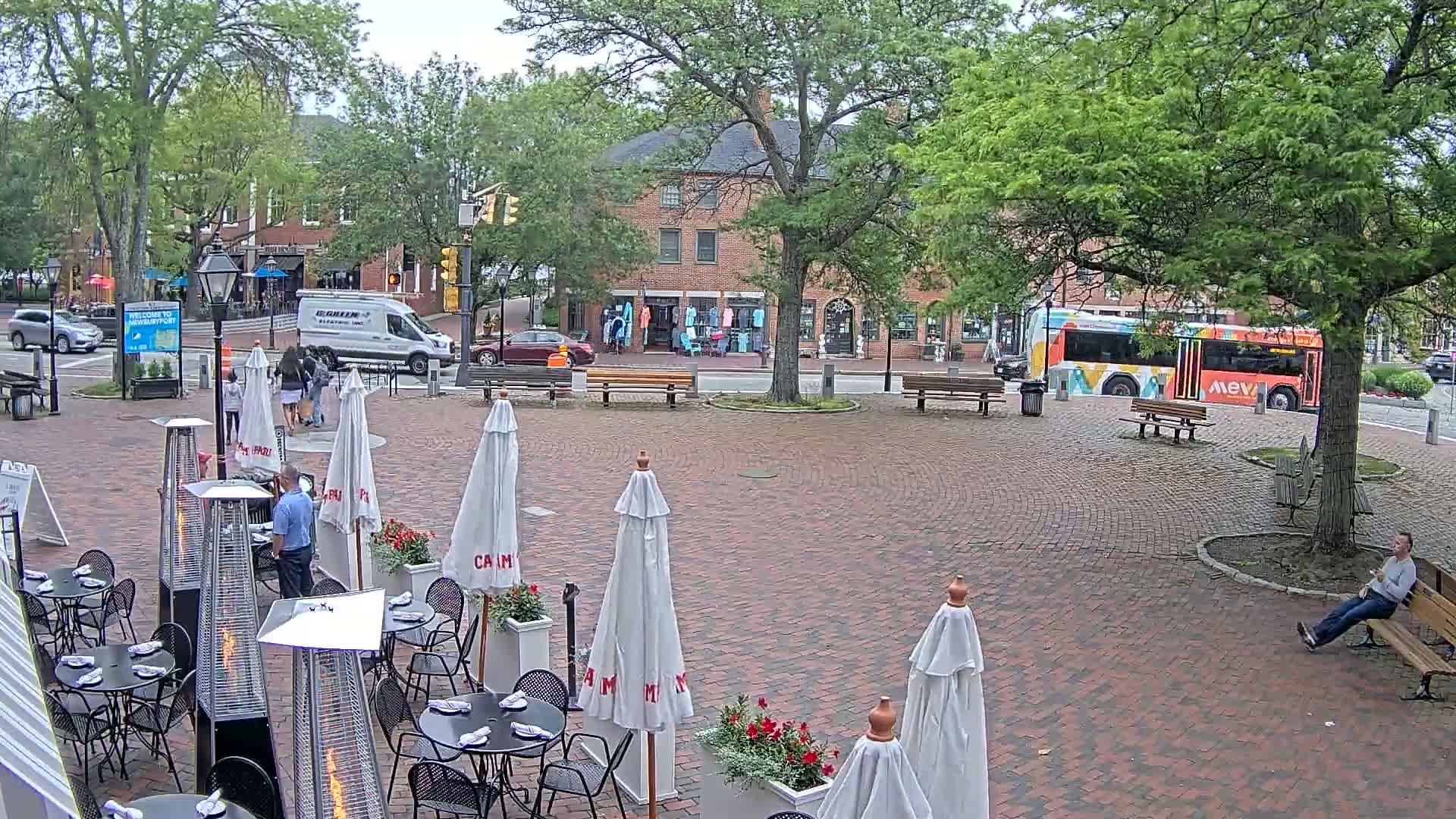 A brick-paved outdoor patio with white umbrellas, patio heaters, and tables and chairs sits under a partly cloudy sky in front of brick buildings, with a bus and some pedestrians visible in the background.