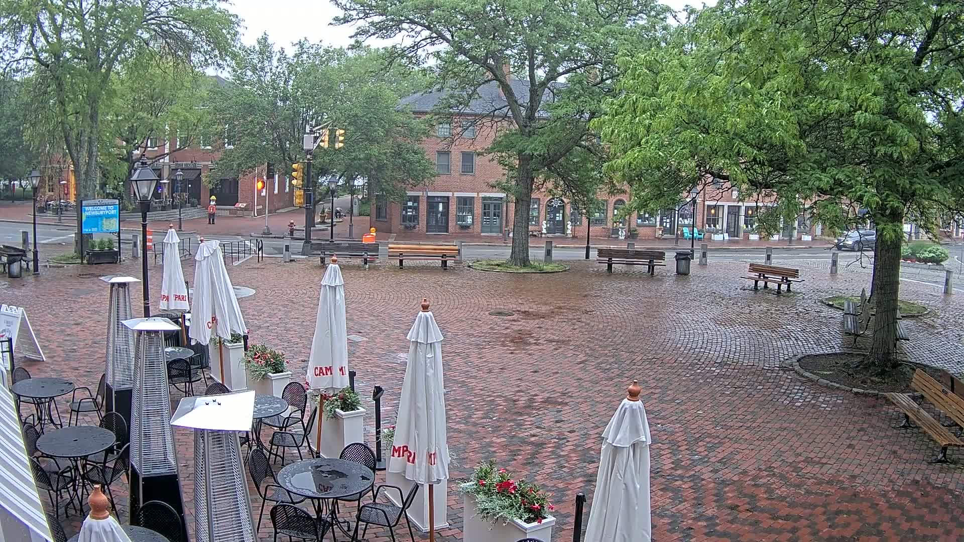 A brick plaza with outdoor cafe seating, umbrellas, and patio heaters, situated in front of brick buildings under an overcast sky.