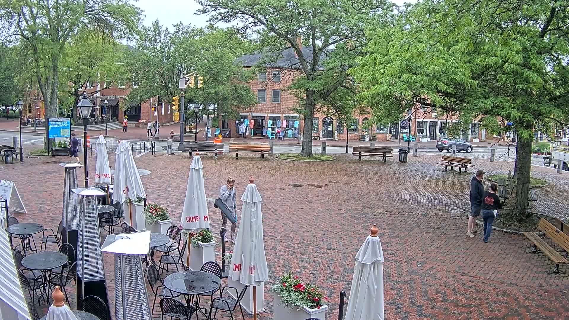A brick plaza with outdoor café seating, surrounded by shops and trees, on a wet day.