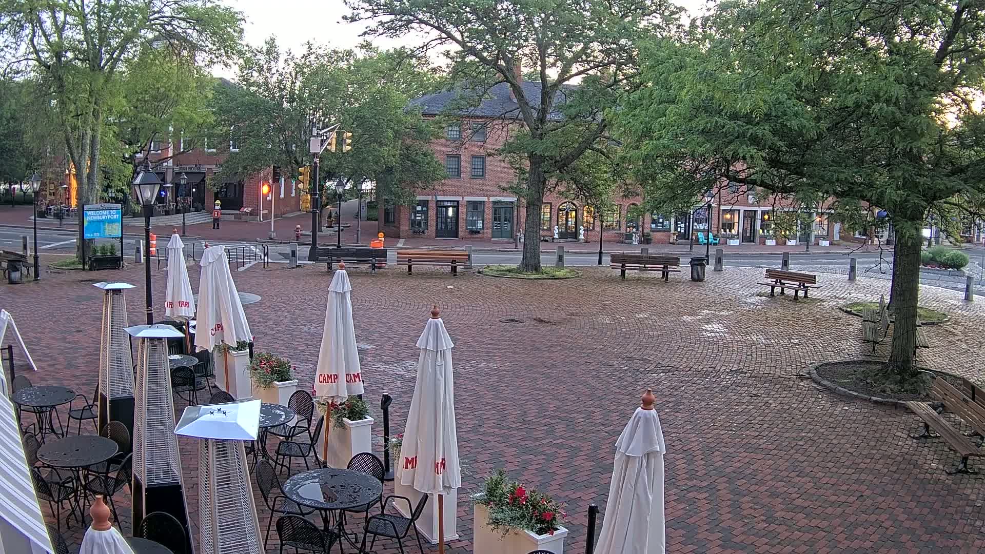 A brick plaza with outdoor cafe seating,  benches, and trees, under an overcast sky.
