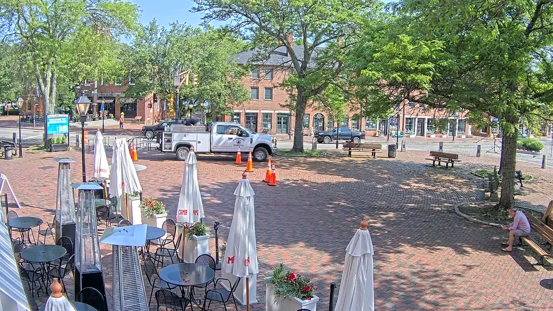 A brick plaza with outdoor seating, patio heaters, and white umbrellas under a sunny sky, surrounded by brick buildings and lush green trees.
