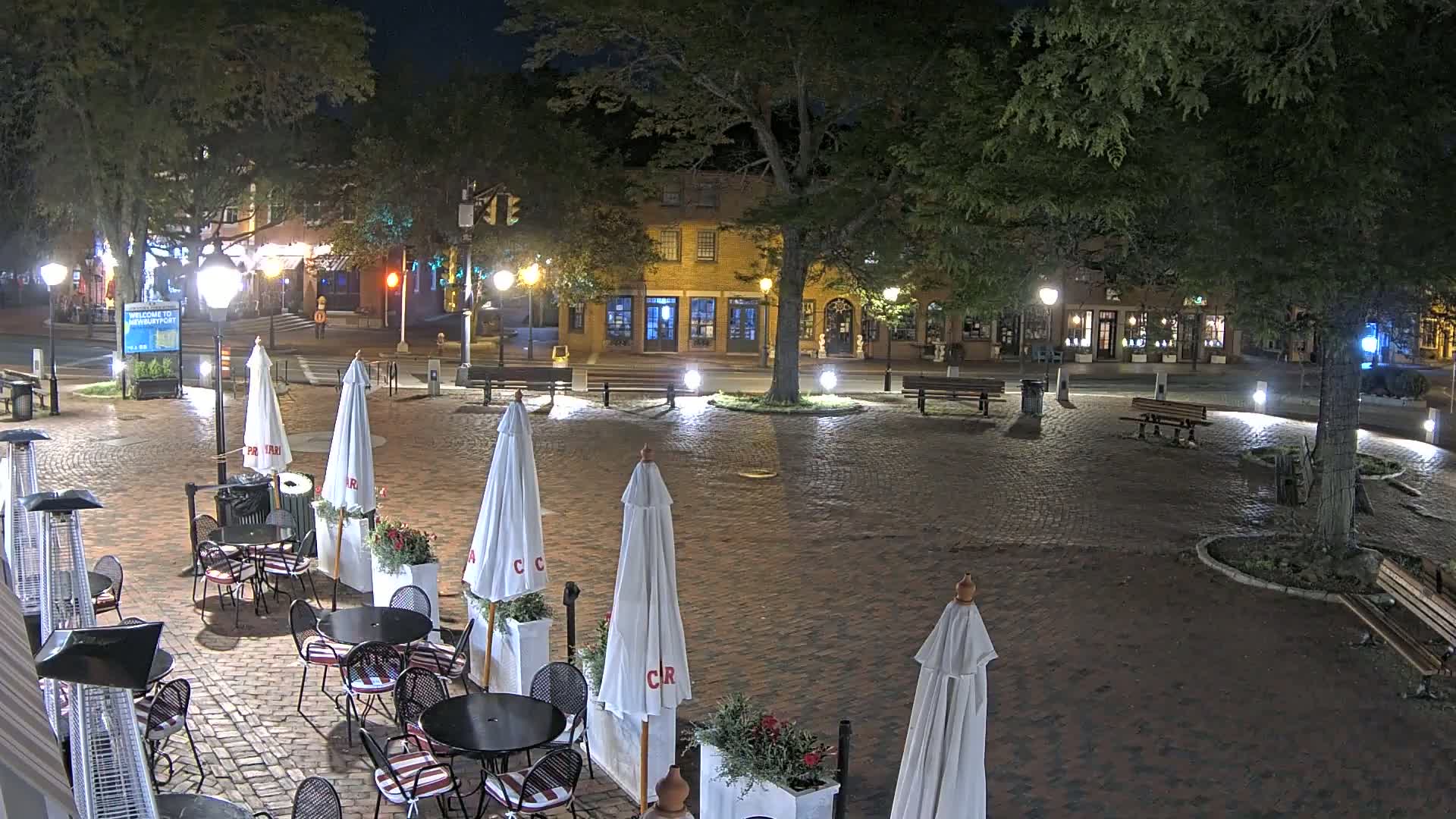 A brick plaza at night, illuminated by streetlights, contains several outdoor cafe tables with umbrellas, benches, and a large tree in the center, surrounded by low-lit buildings.