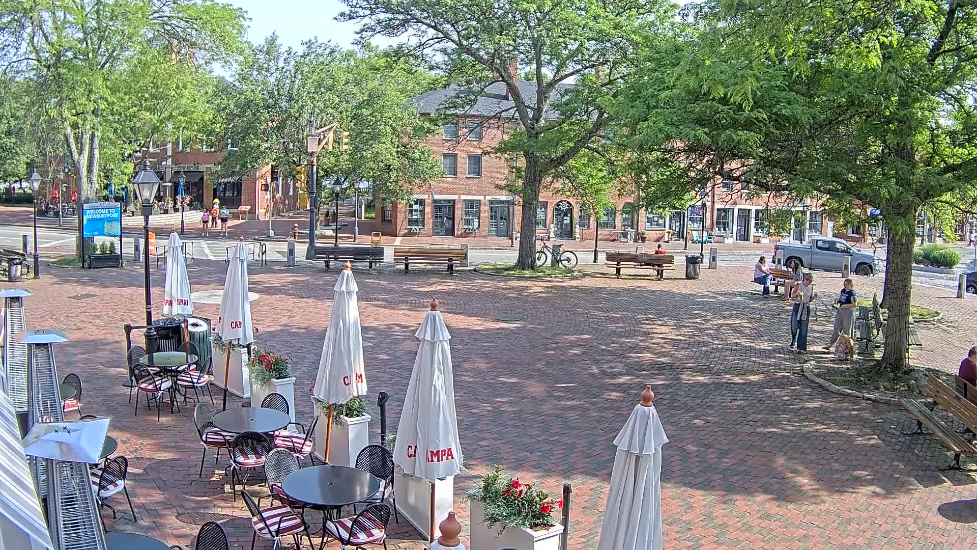 A brick plaza with outdoor cafe seating, shaded by umbrellas and trees, on a sunny day in front of brick buildings.