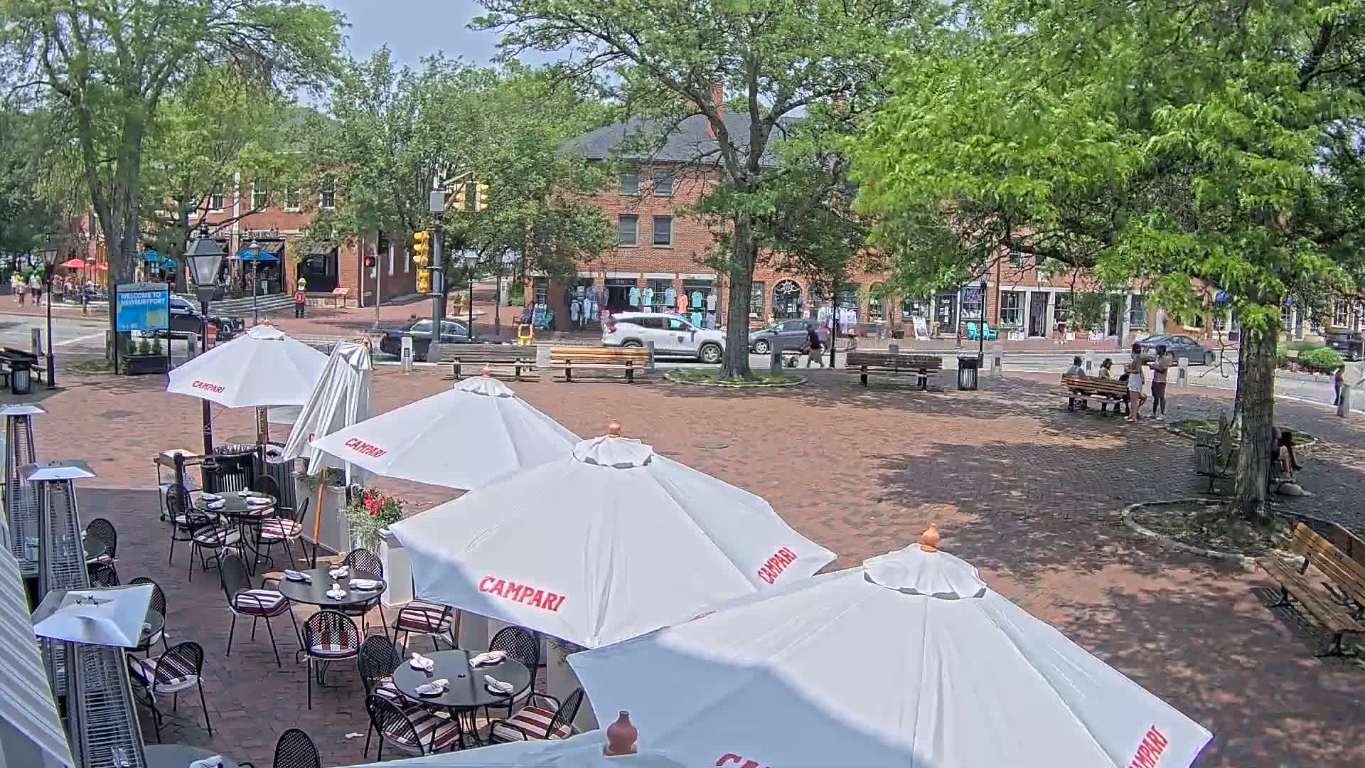 An outdoor brick plaza with several patio umbrellas and tables, surrounded by buildings and trees on a sunny day.