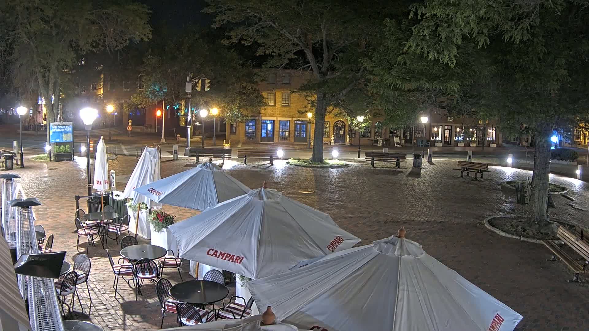A nighttime view of a brick plaza with outdoor cafe seating under several umbrellas, surrounded by trees and buildings with illuminated windows.